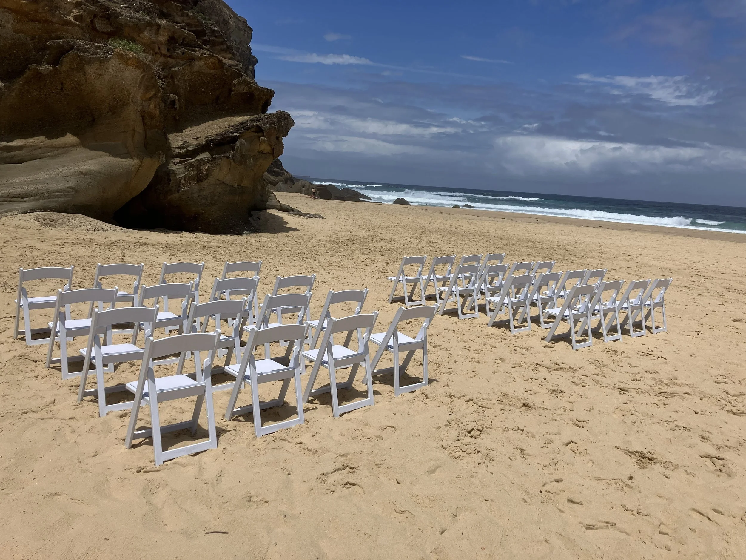 Empty white chairs arranged in rows on a sandy beach facing the ocean, with a large rock formation on the left and a cloudy sky overhead.
