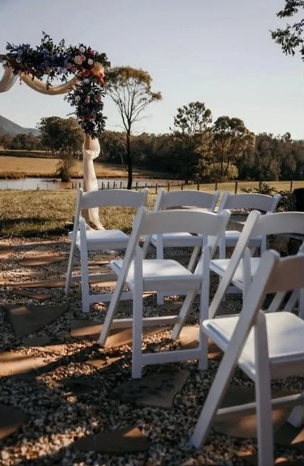 Outdoor wedding ceremony setup with white chairs on gravel, floral arch, and scenic landscape with trees and water in the background.