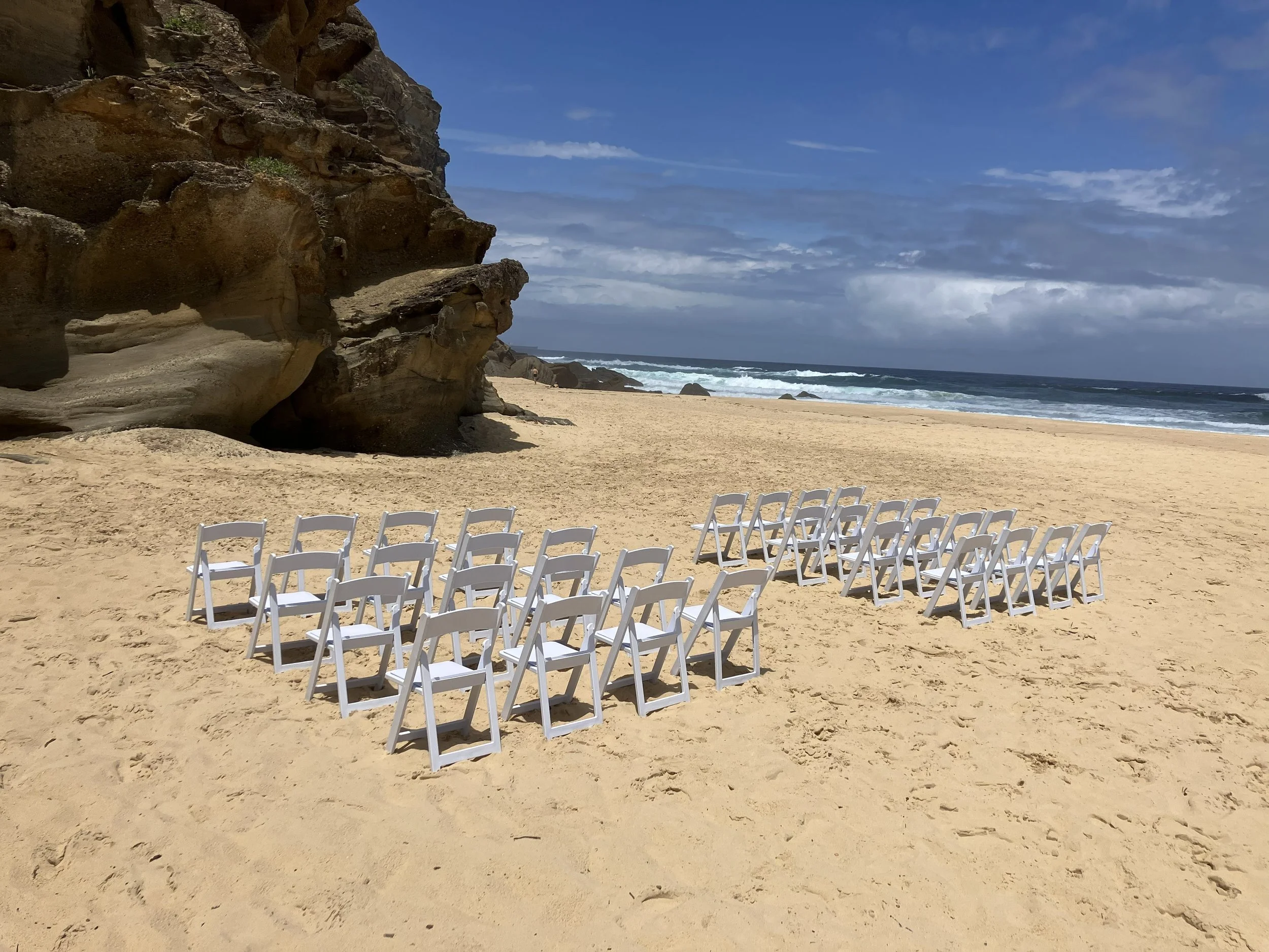 Empty white chairs arranged on a sandy beach facing the ocean with waves, rocky cliffs on the left, and a blue sky with clouds.