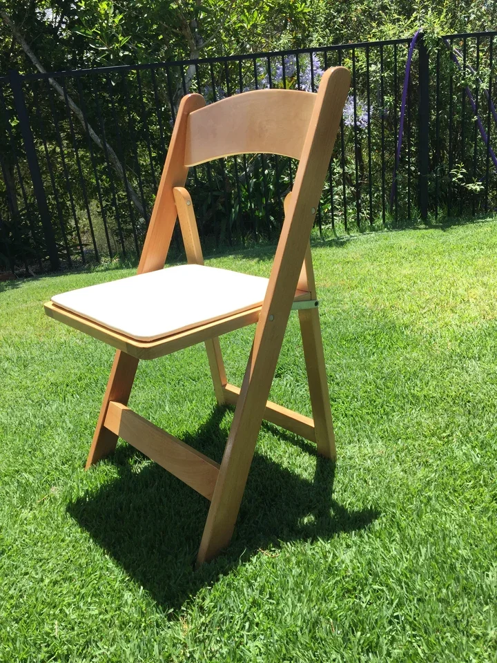A wooden high chair with a beige backrest and a white seat cushion, placed on a green lawn with a black metal fence and trees in the background.