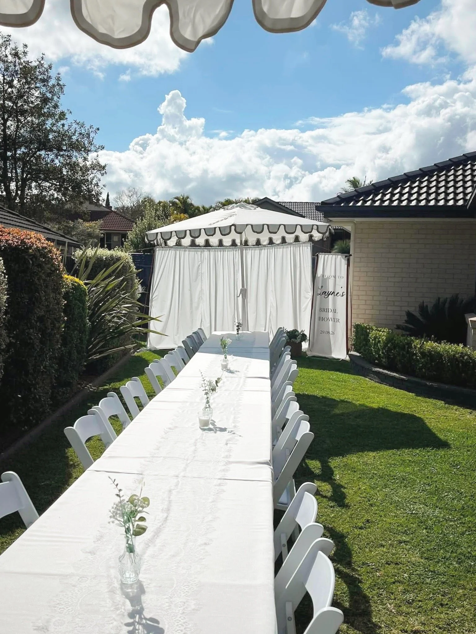 Outdoor garden setting for a wedding or event with a long white dining table decorated with small flower vases, surrounded by white chairs, with a white canopy in the background, a sign reading 'Welcome to Layne's Bridal Shower,' and a bright blue sk