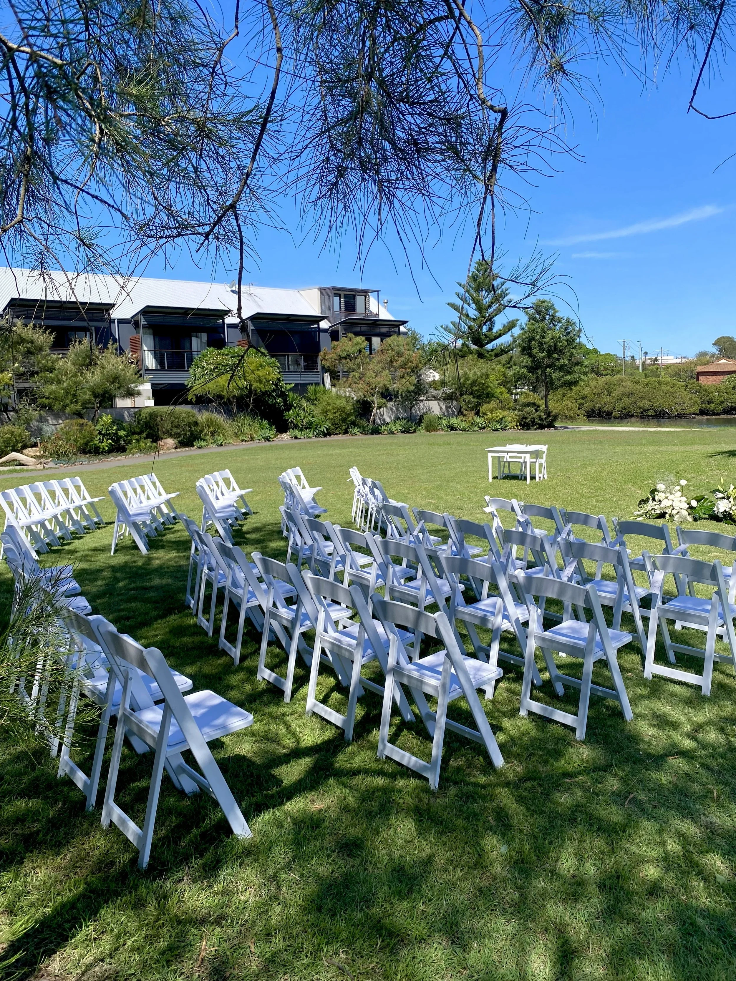 Outdoor wedding setup with white chairs arranged in rows on a green lawn, and a small white table nearby, under trees with a modern building in the background on a clear, sunny day.