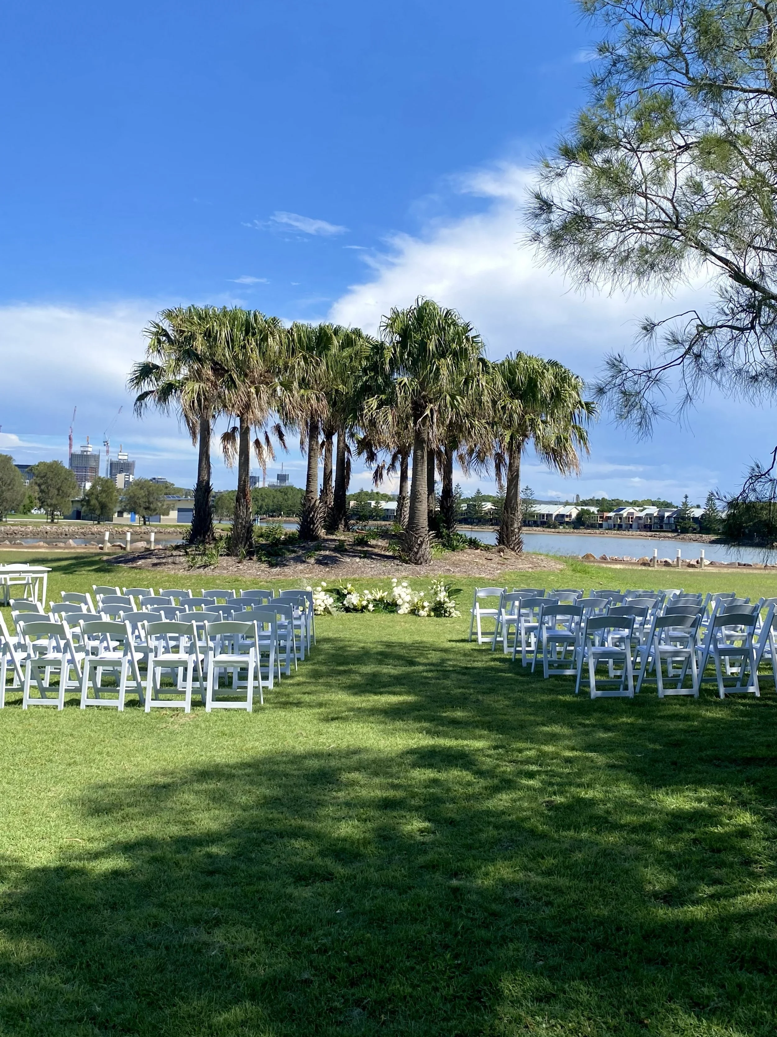 Outdoor wedding ceremony setup with white chairs arranged in rows on green grass, near a small pond, surrounded by palm trees and other greenery, with a cityscape with buildings in the background under a partly cloudy sky.