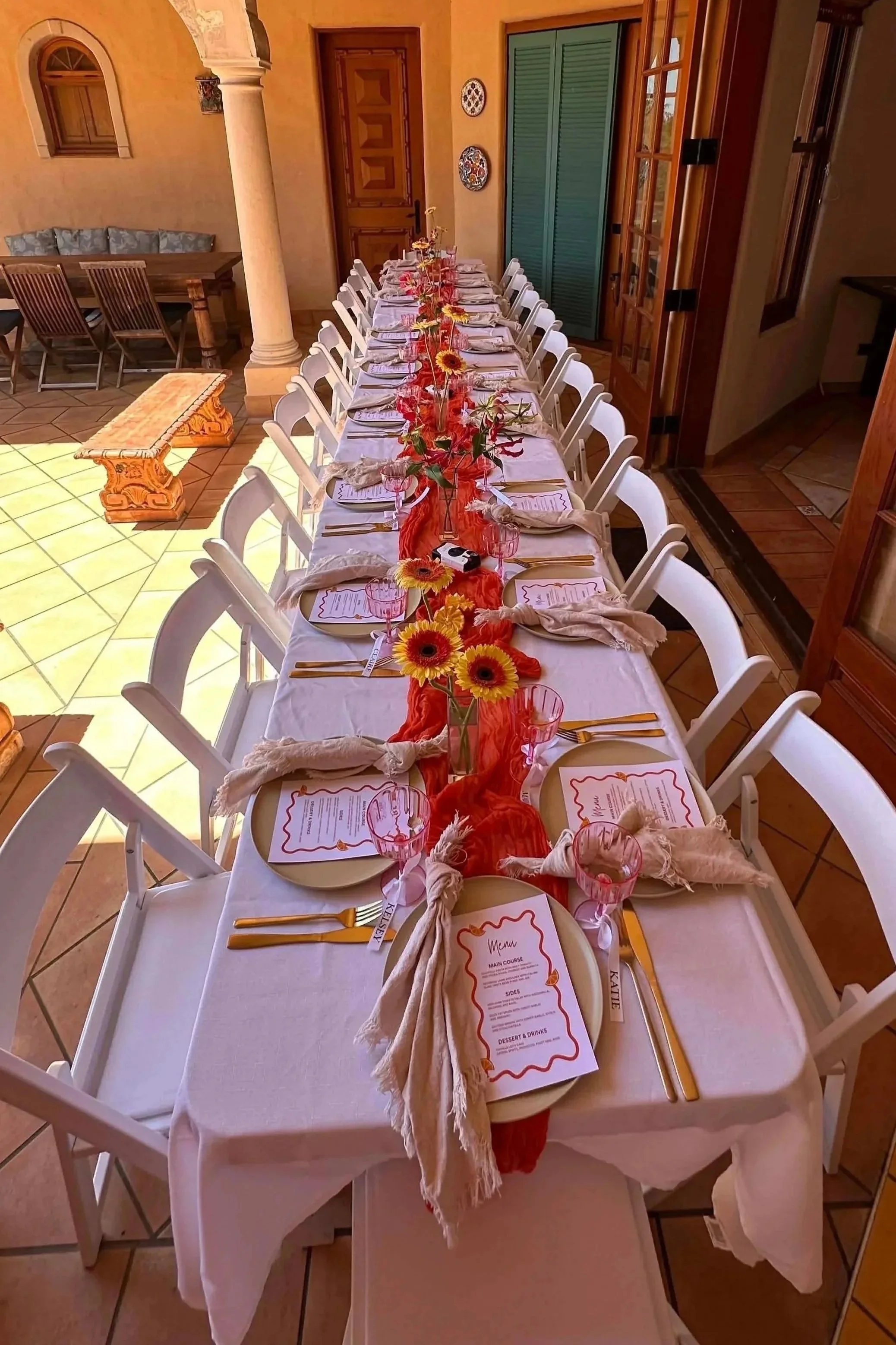A long dining table set up for a meal with white chairs, beige napkins, gold utensils, pink geometric glasses, and sunflower centerpieces on an orange table runner. The scene is in an indoor patio with terracotta tiled flooring, decorative wall plates, and a background of dining area with wooden furniture and wooden doors.