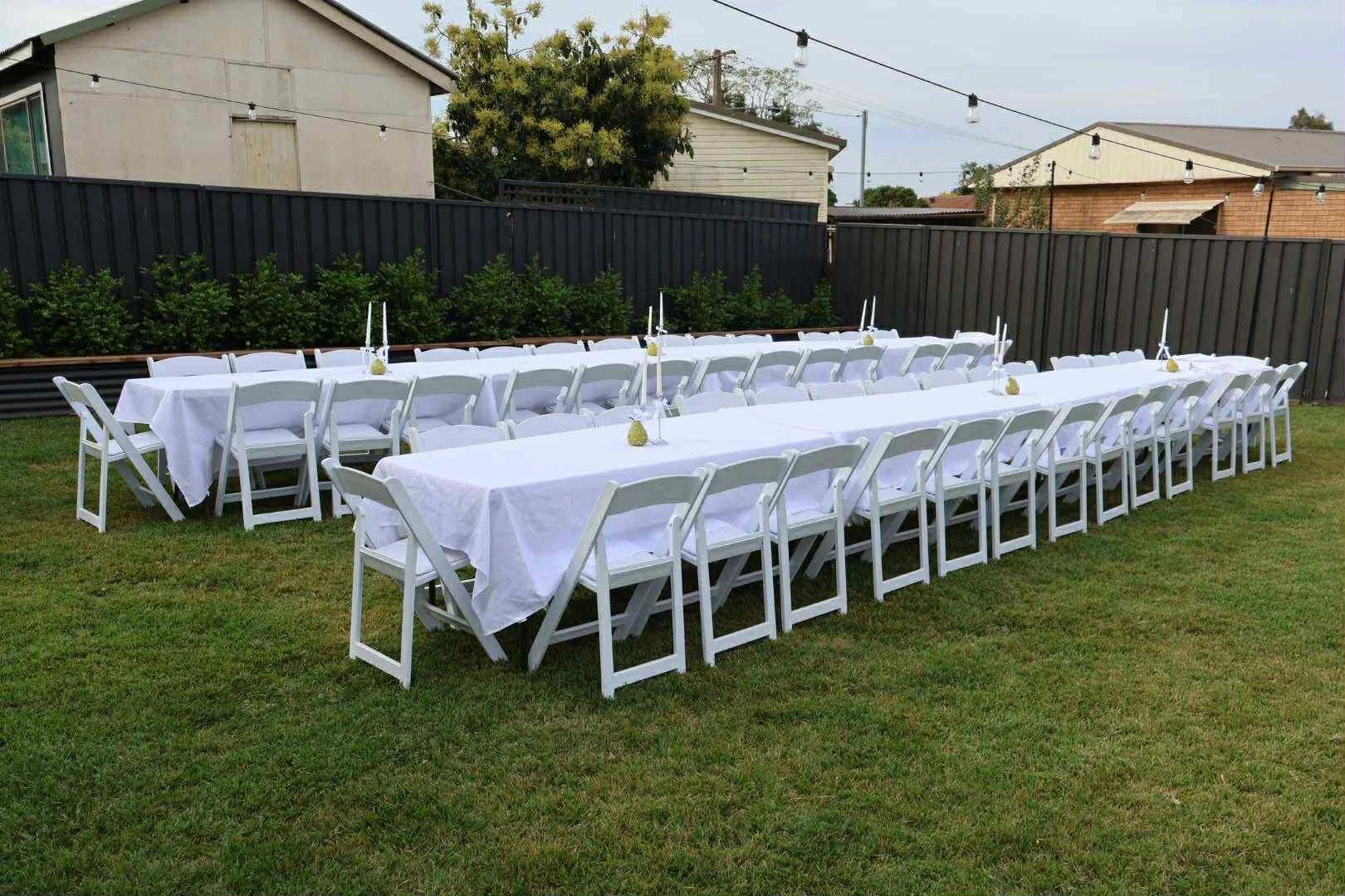 White rectangular tables with white folding chairs arranged outdoors on grass, decorated with white tablecloths and small centerpieces, set for a gathering or event under string lights, with a wooden and brick fence and houses in the background.