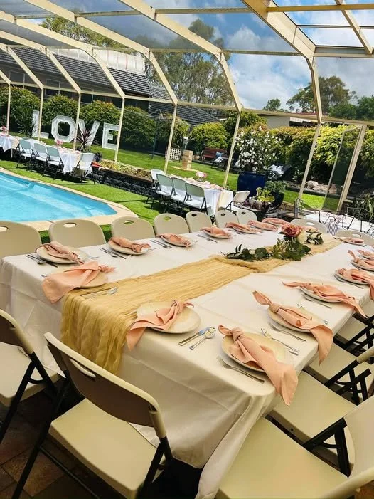 Long dining table set for a celebration with pink napkins and a floral centerpiece, located outdoors under a screened enclosure with a pool and greenery in the background.