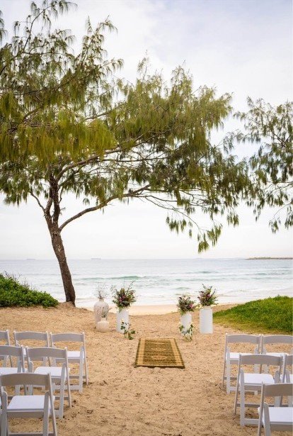 Beachside wedding setup with white chairs, floral arrangements, and a tree overlooking the ocean.