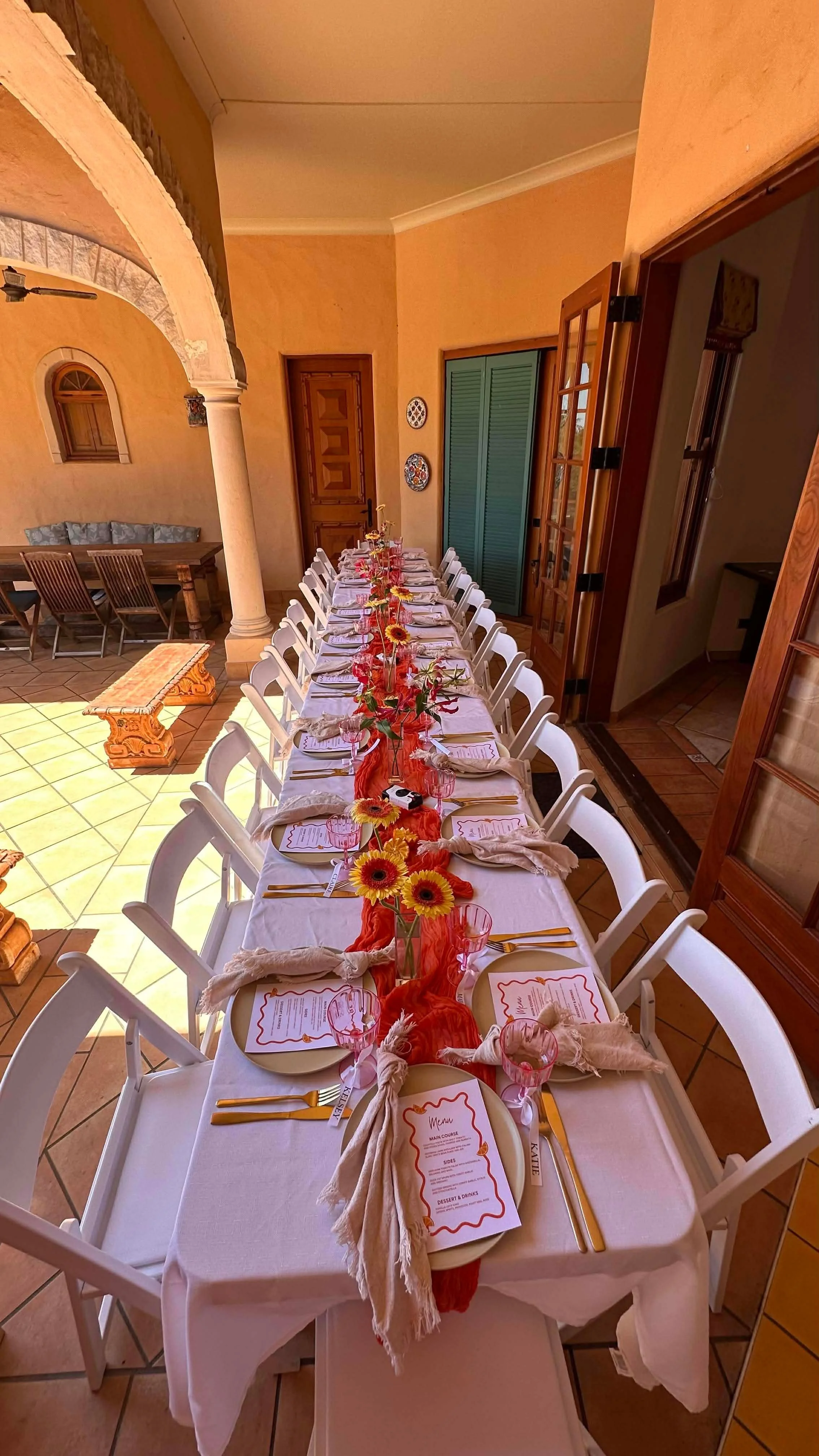 Dining table set for a celebration with floral centerpieces, table settings, and menus in a warm-colored room with arched doorways and tiles.