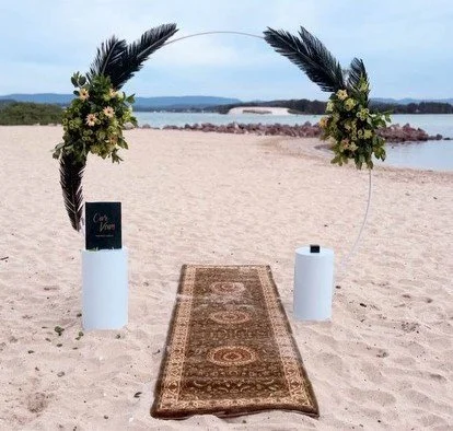 Beach wedding arch with floral arrangements, palm leaves, and a patterned rug on sand, with water and distant land in the background.