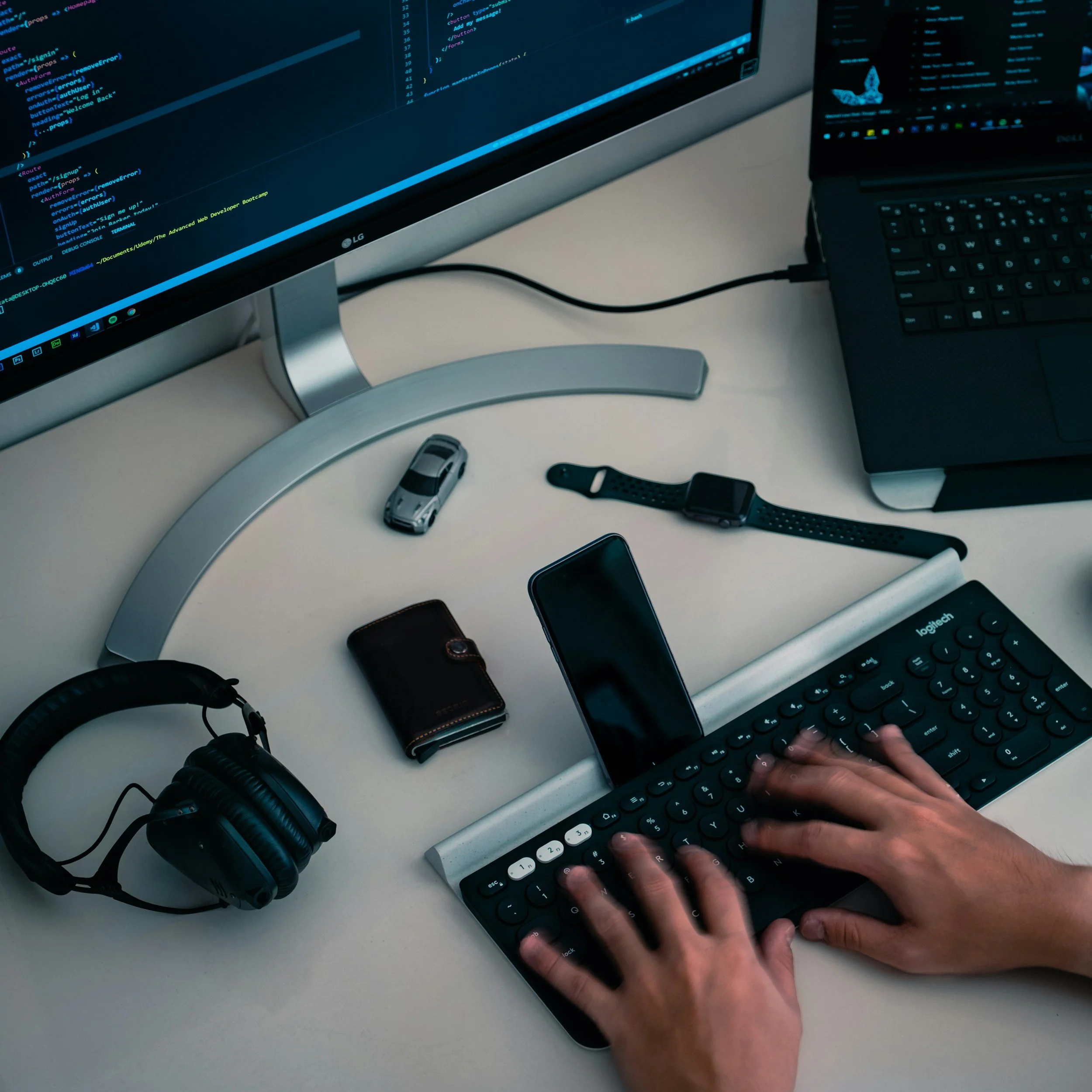 A person is typing on a computer keyboard with code displayed on an LG monitor, alongside a laptop, smartphone, and various accessories on a white desk, with a pair of headphones and a small toy car also visible.