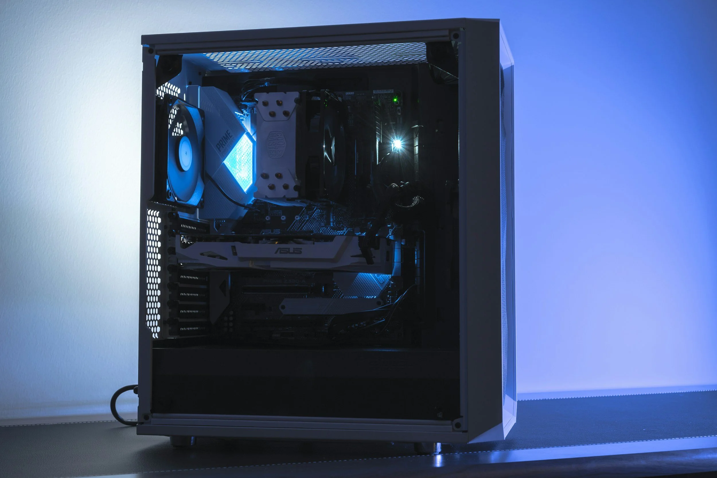 A gaming computer with a transparent side panel showing internal components, illuminated with blue LED lights, placed on a desk against a blue-lit background.