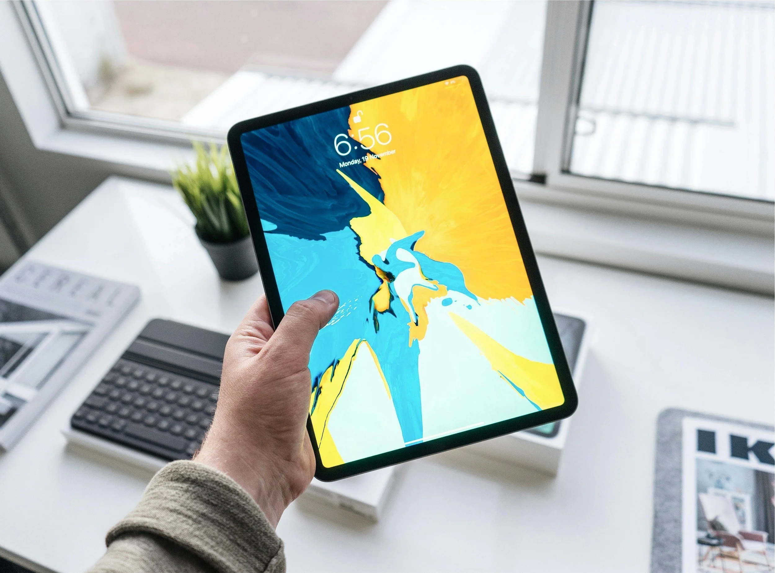 Person holding a tablet displaying a bright abstract colorful wallpaper, in a workspace with a computer keyboard and monitor, and a window with a view outside.