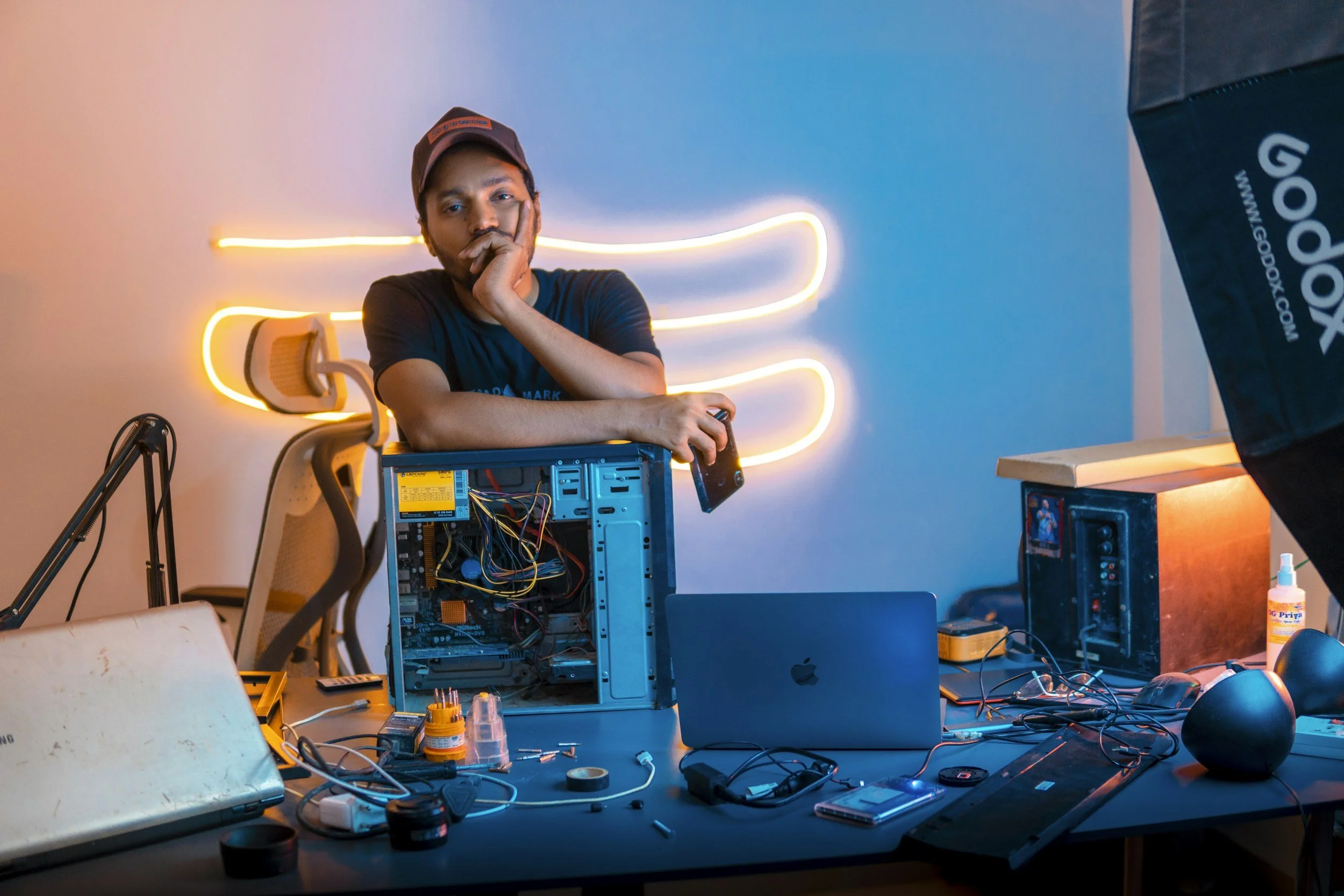 A man with a hat and black shirt sits at a cluttered table with electronics tools, wires, and a computer, with a neon light behind him in a room with a blue wall.