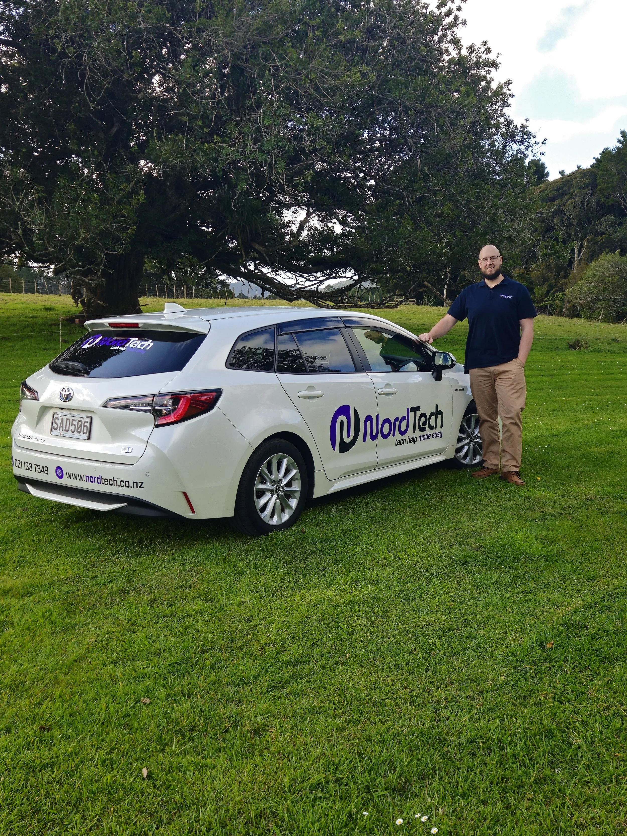 A man standing next to a white car with NordTech branding in a grassy field with large trees in the background.