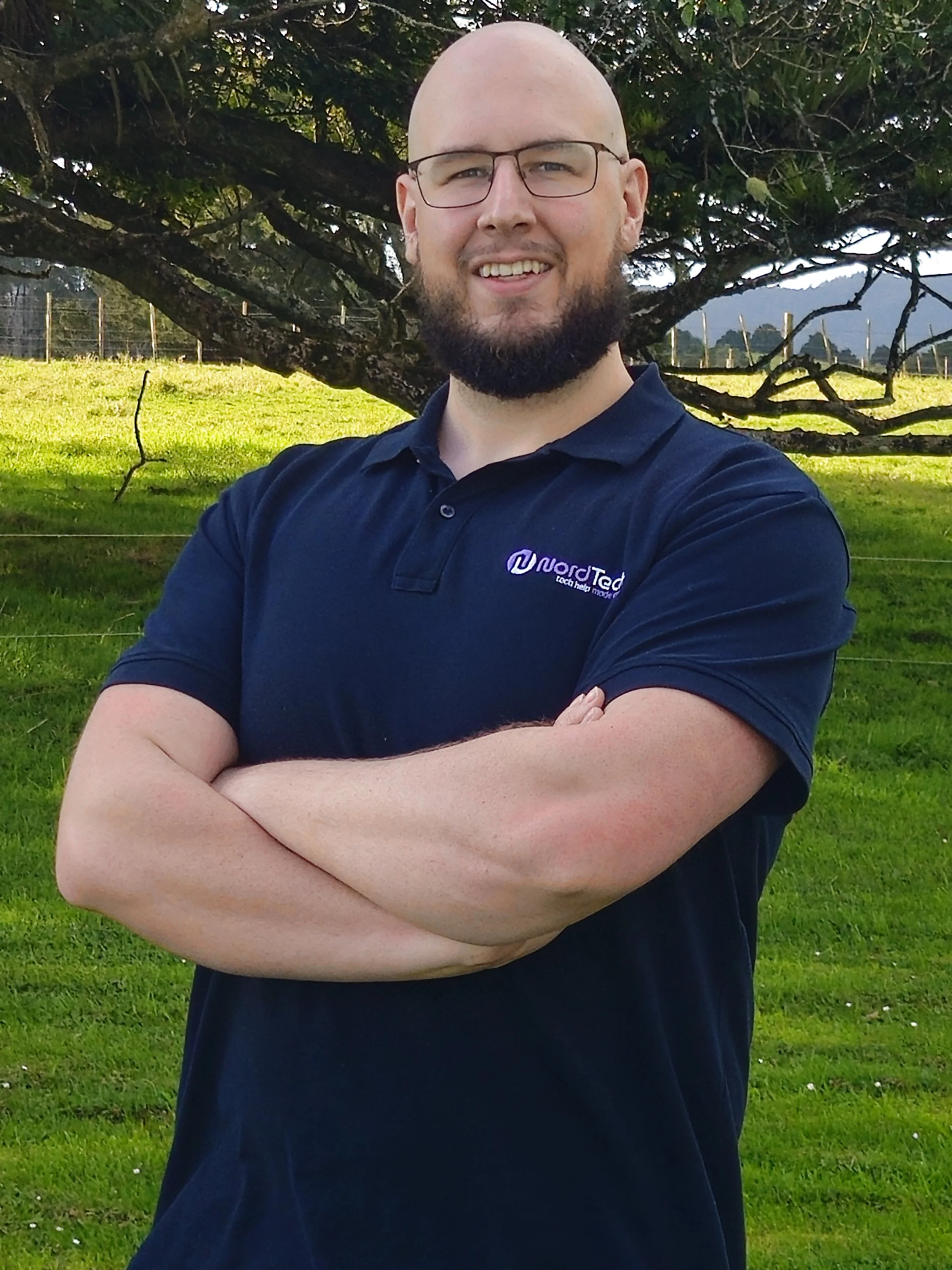 A bald man with glasses and a beard smiling, standing outdoors in front of a tree with green grass and a distant fenced area.
