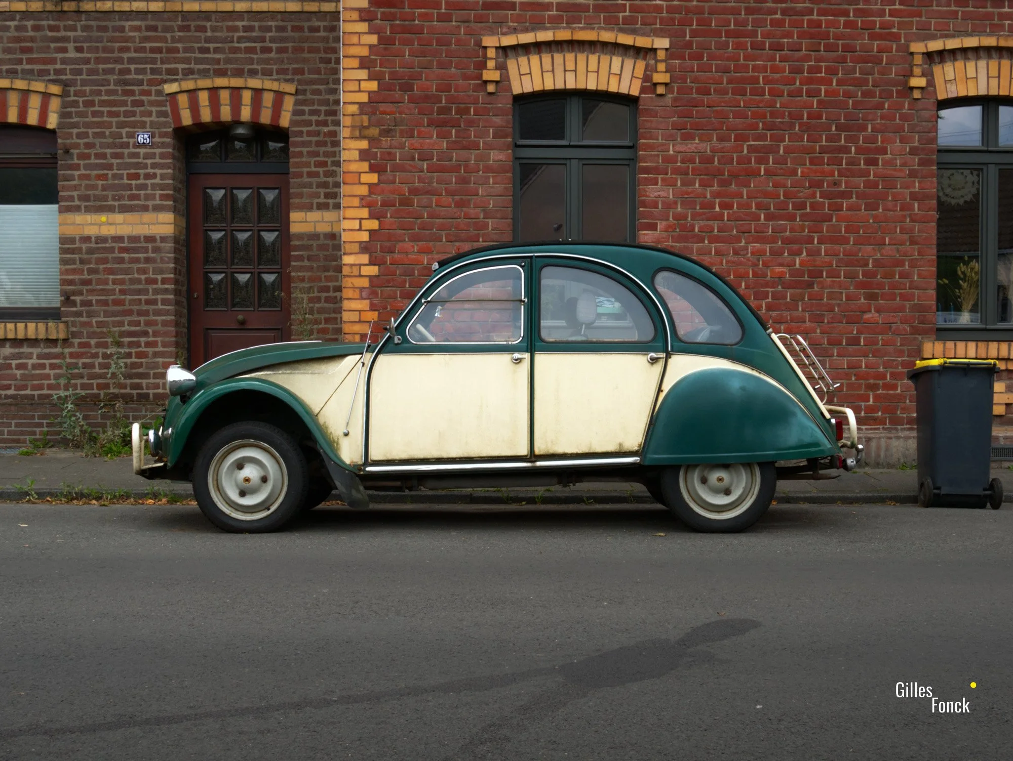 Green Citroen 2CV in side profile in front of a red brick facade. A trash can is neatly placed to the right of the car.