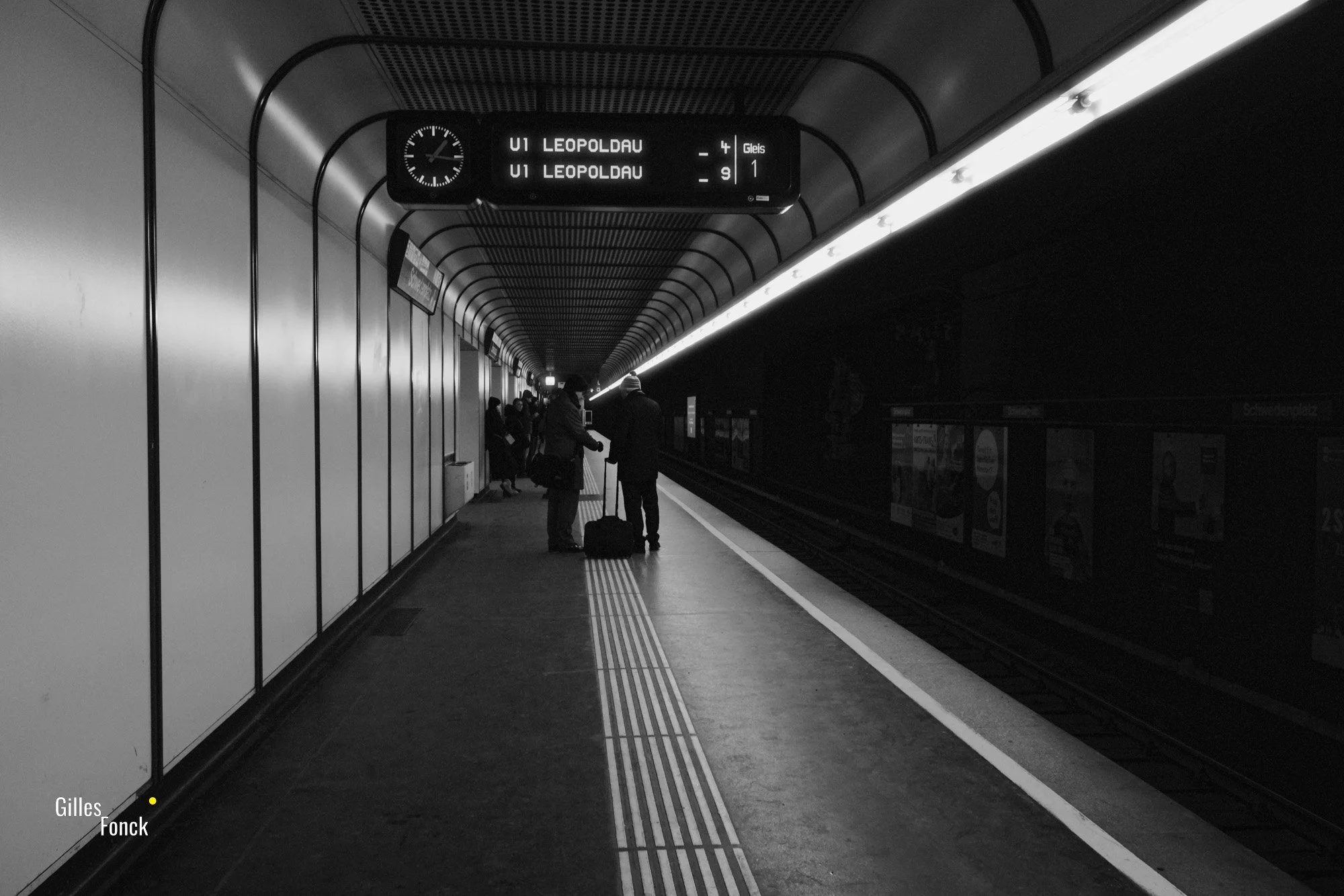 Monochrome picture of a platform at a70s style underground station in Vienna. The lines of the platform are leading into the distance, people are waiting for a train.