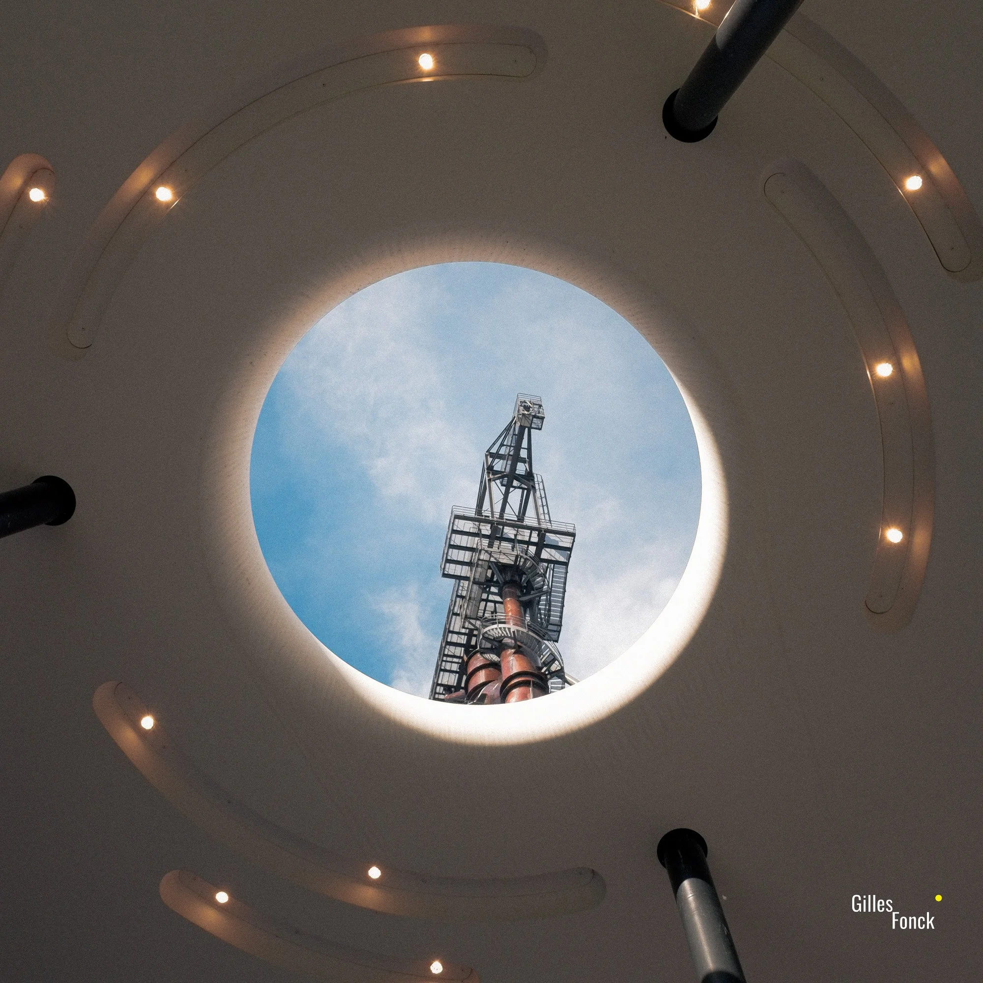 The tower of a blast furnace can be seen through a round opening in the roof of a shelter. LED spots are set into the roof.