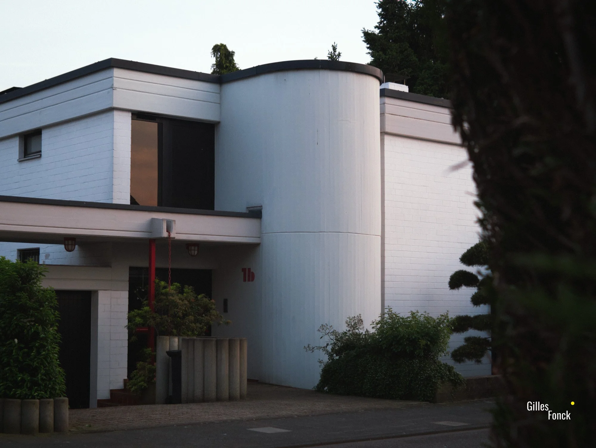 A modernist home in the suburbs of Colgone. Built of white bricks and concrete with a flat roof, it's house number and some structural details are accented in a red color.