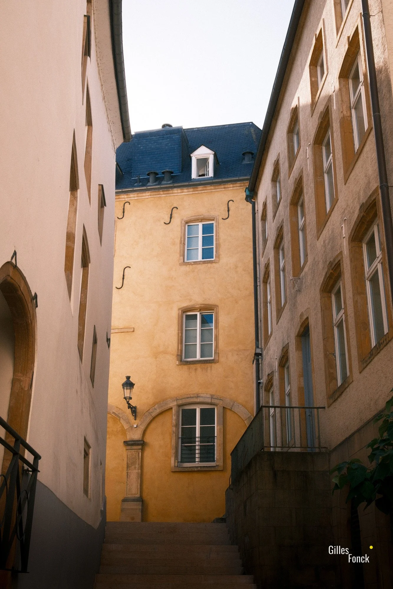 A small flight of stairs between historic buildings leeds up to a yellow facade with an old street lamp. The colors and washed out look of the facade almost makes ist look painted.