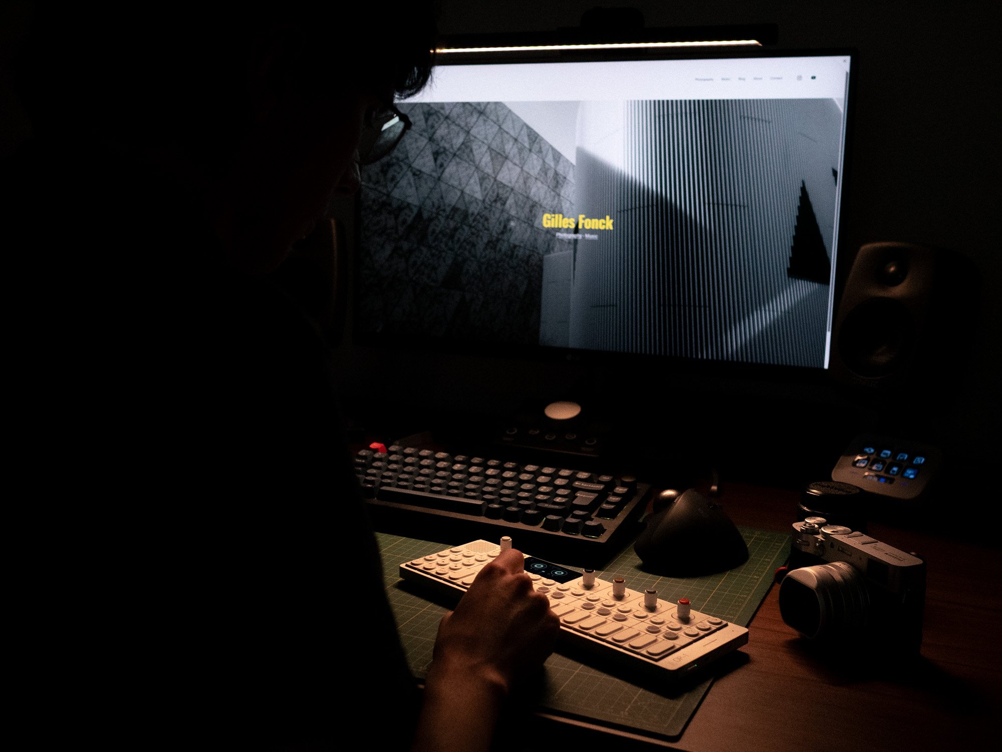A person working at a desk with computer equipment in a dark room. The person is operating a LEGO control panel with various knobs, and there is a lit computer monitor displaying an architecture or design program.
