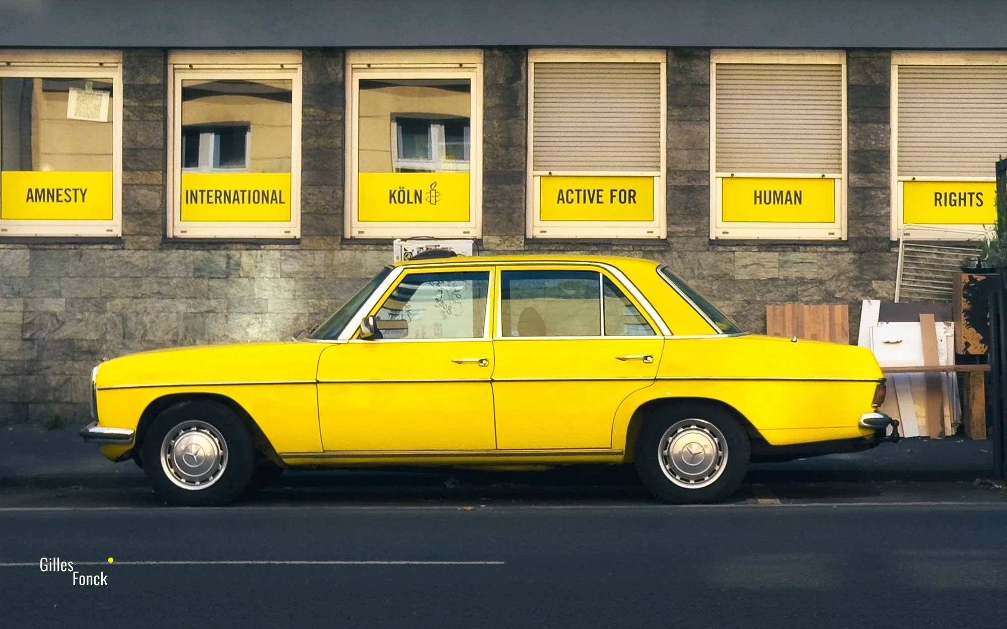 Yellow old limousine in side profile in front of windows covered in yellow advertisements for Amnesty International