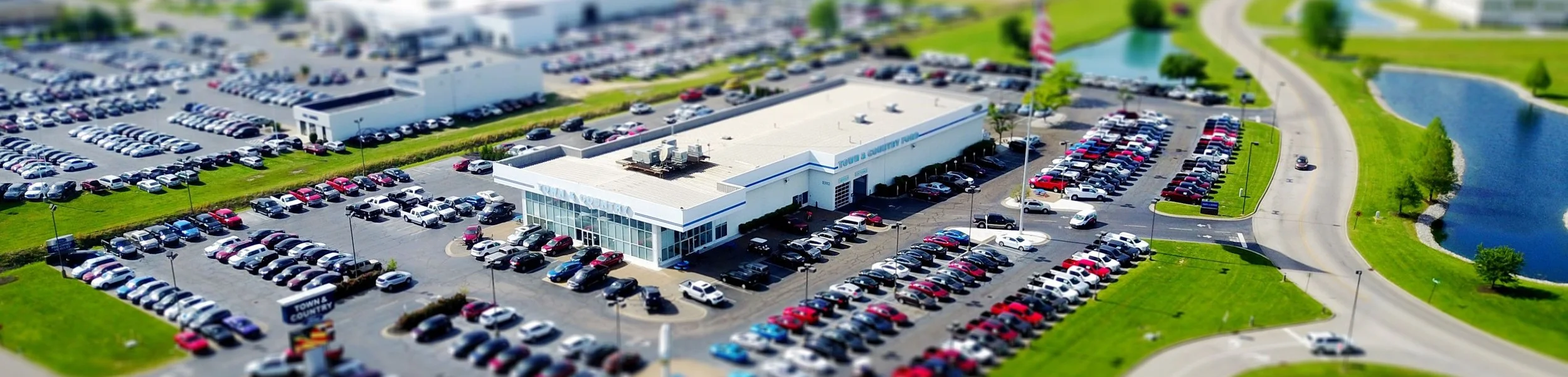 Aerial view of a town hall building with a large parking lot filled with cars, on a sunny day with green grass, trees, and a nearby pond.
