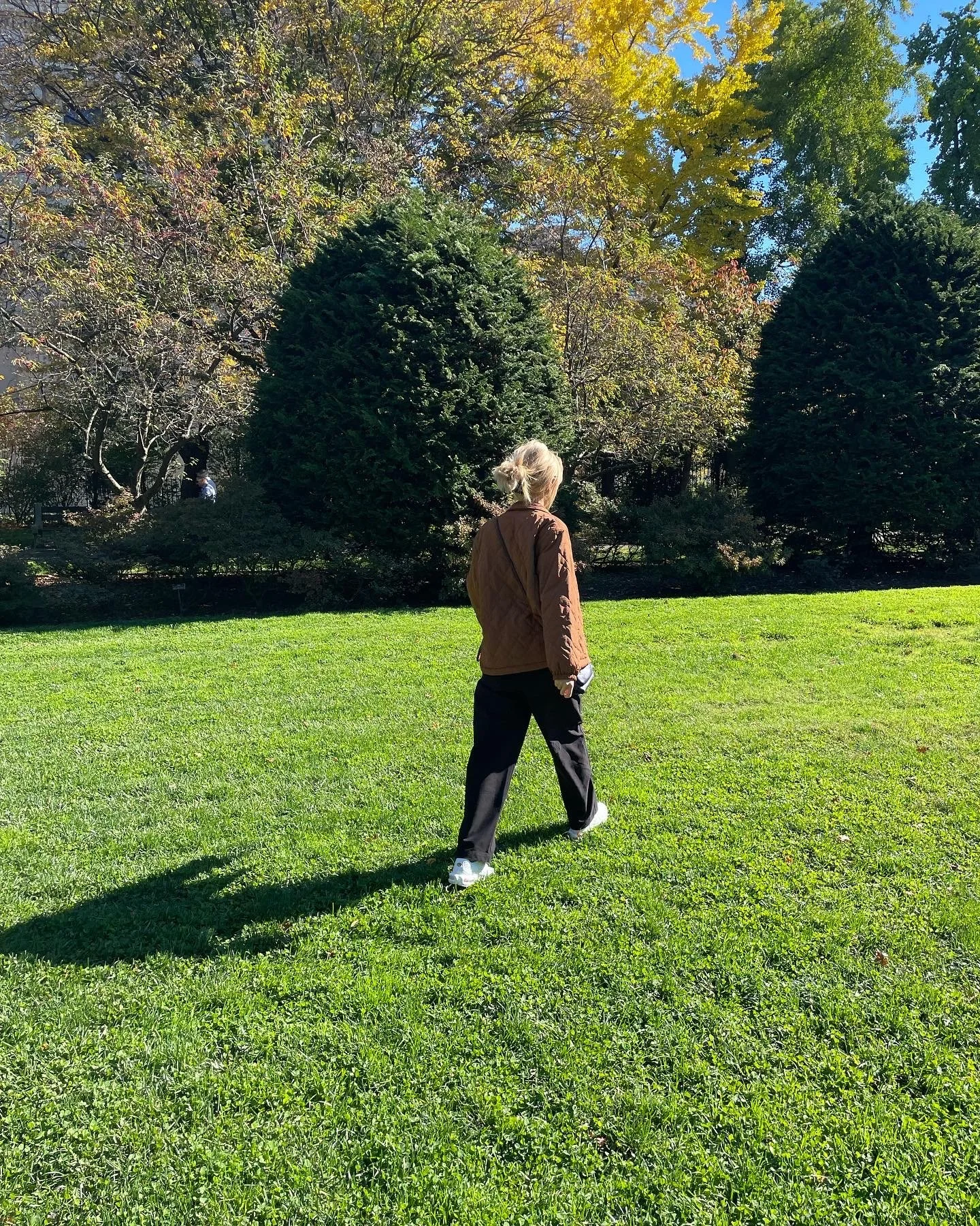 A woman walking on a grassy lawn in a park or garden during autumn, with trees in the background showing yellow and green leaves on a sunny day.