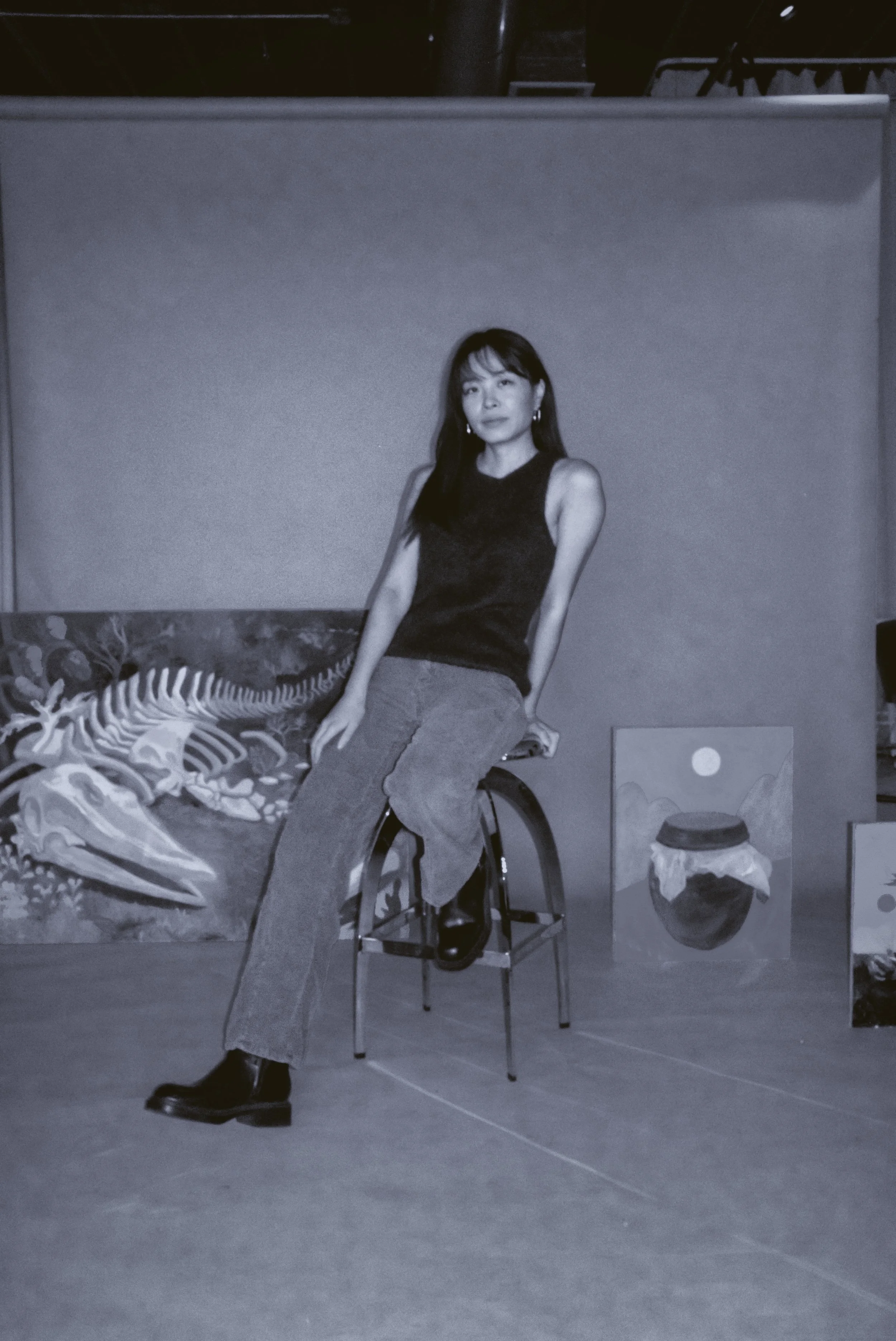 A woman with long dark hair, wearing a sleeveless black top, light-colored jeans, and black ankle boots, sits on a chair in an art gallery or studio setting. She is looking at the camera with a neutral expression. There are paintings and artworks in the background, including one with a minimalist landscape, and a table with a cloth covering it.