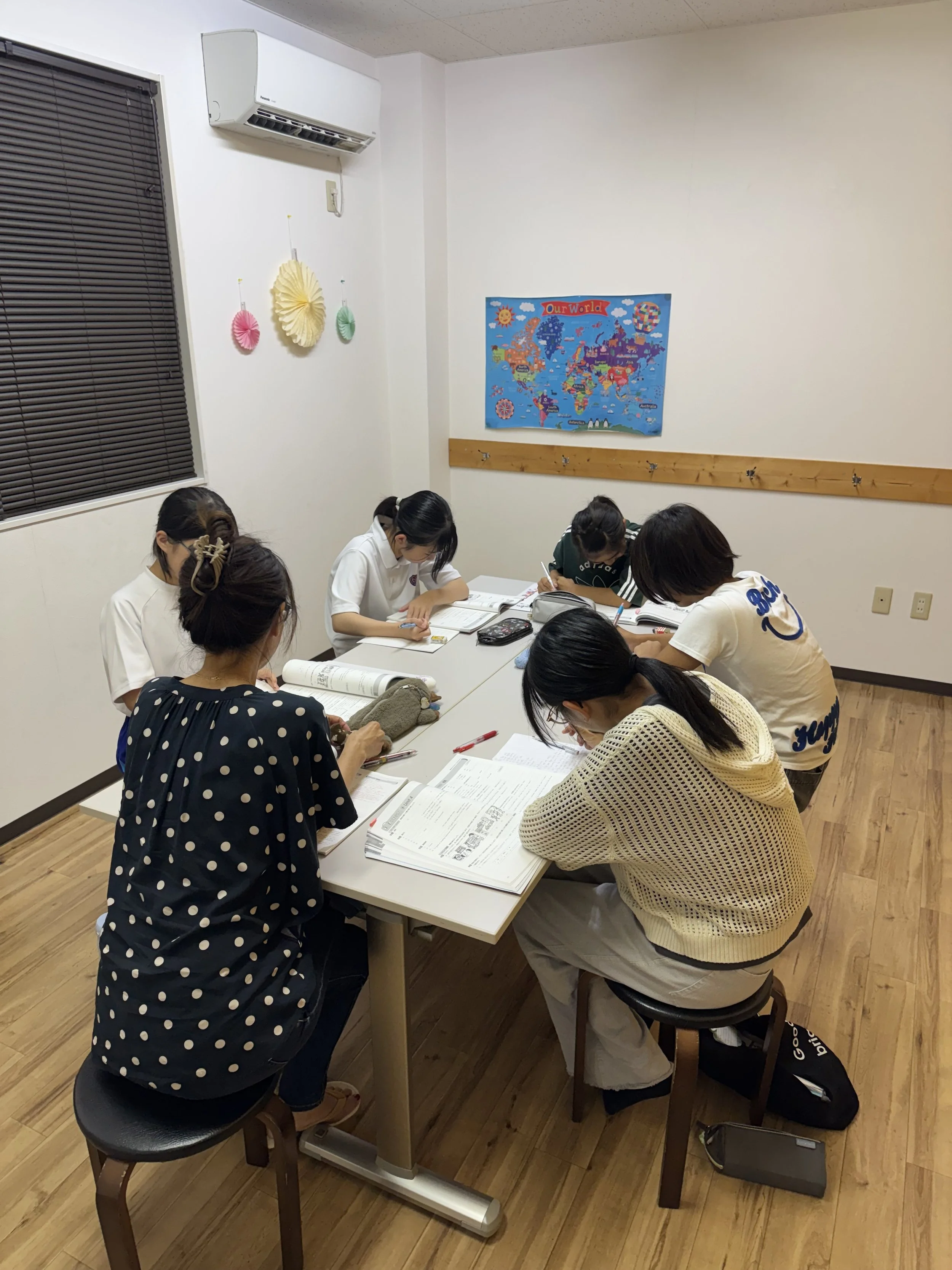教室で複数の生徒がテーブルを囲み、教材を使って学習している様子。壁には世界地図のポスターと飾りがある。