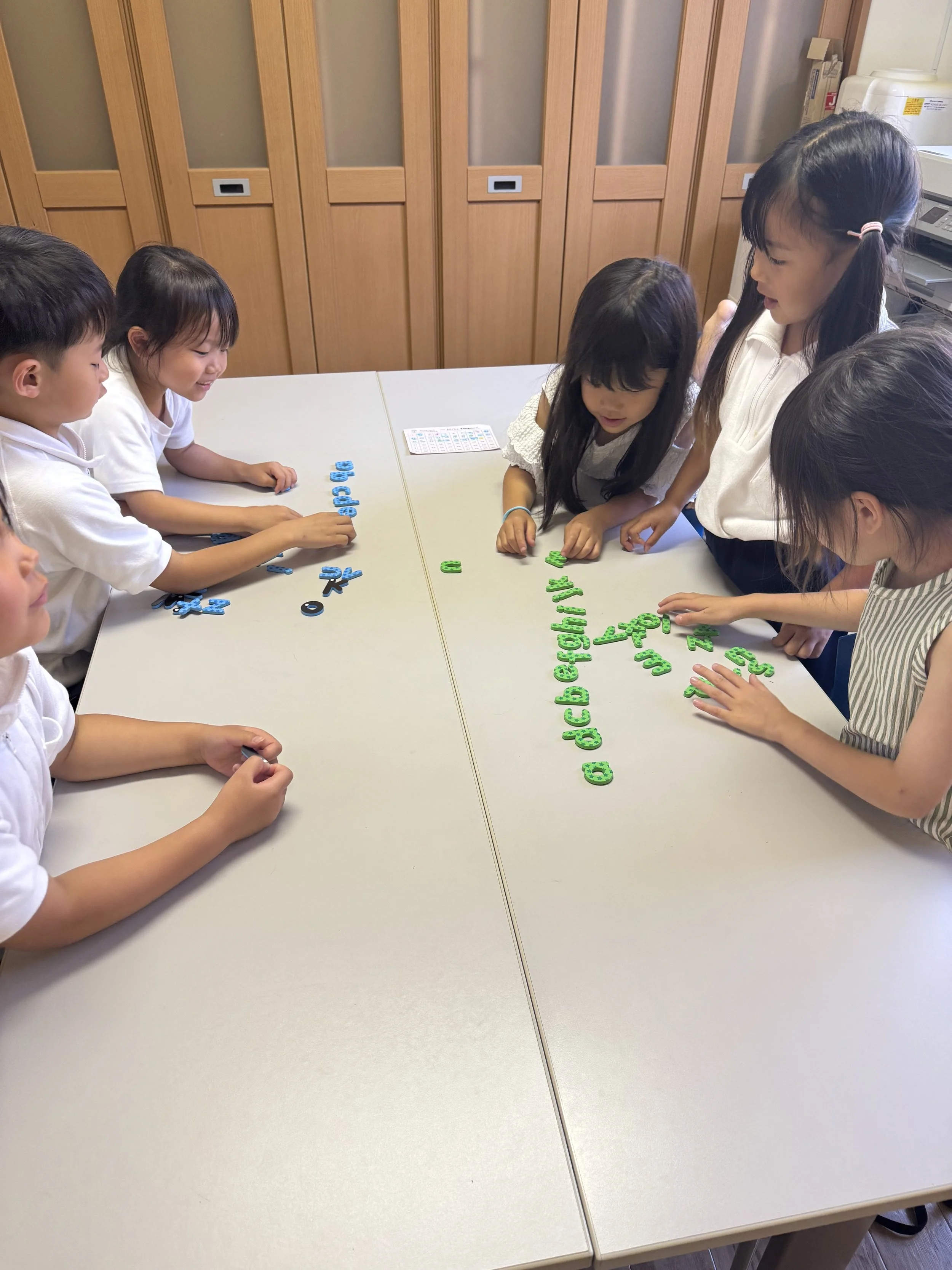 子どもたちと大人がテーブルを囲み、カラフルな文字パーツで遊んでいる様子