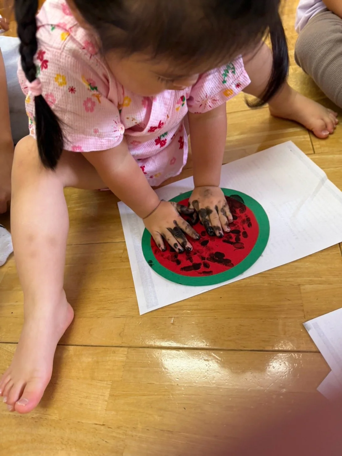 花柄の服を着た幼い女の子が、スイカ型の画用紙に手形を押している様子