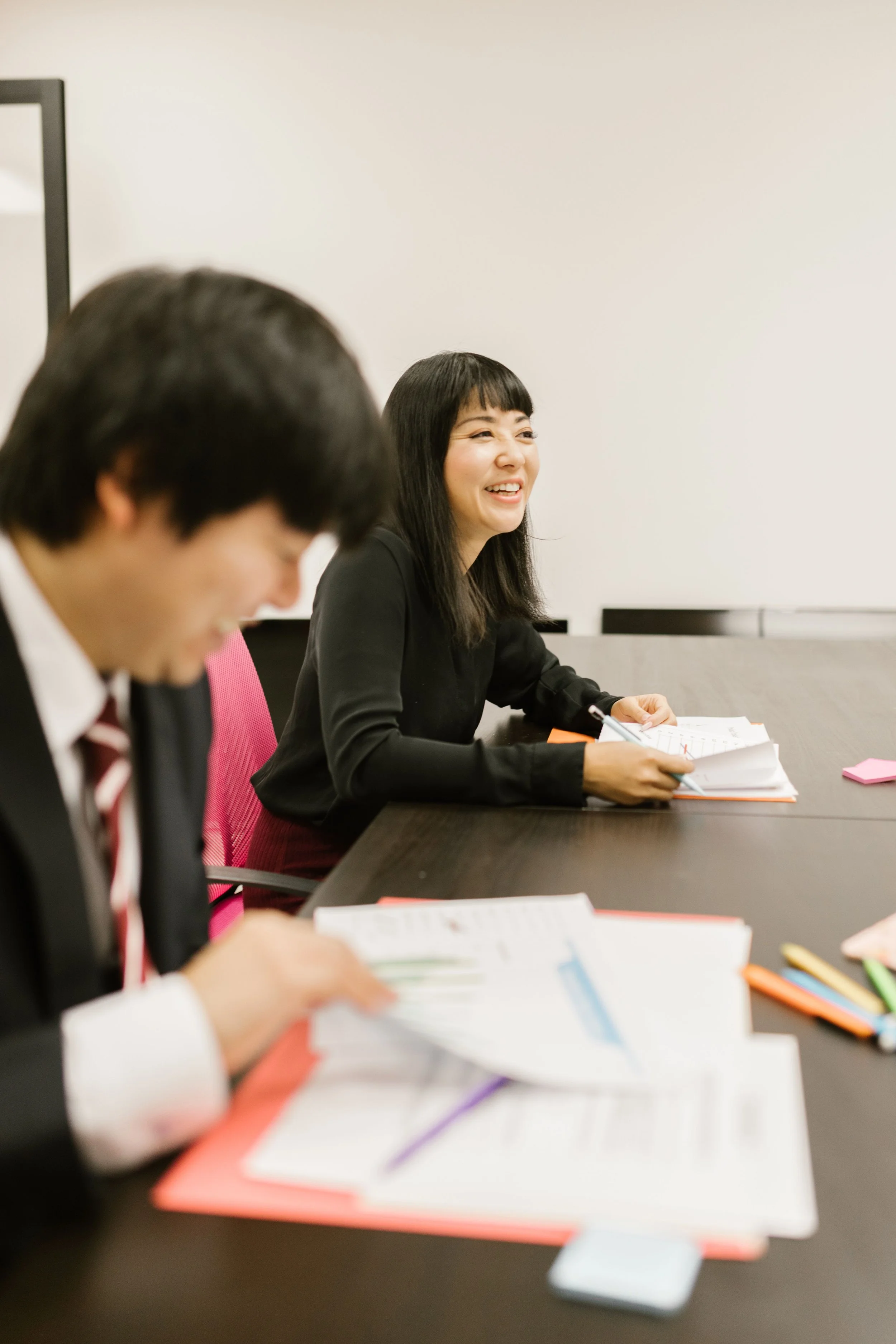 会議室で資料を見ながら笑顔で話す女性と、テーブルを囲む参加者
