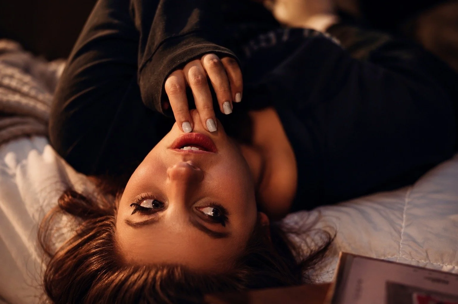 A woman lying on a bed, touching her lips with her fingers, looking at the camera. She has blue eyes, makeup, and brown hair. She's wearing a dark top with shiny nails.