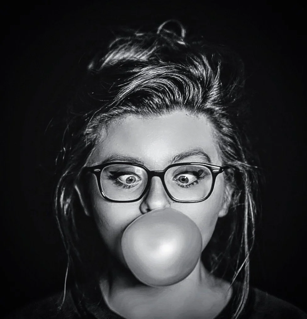 A black and white photo of a woman with glasses blowing a bubble with gum.