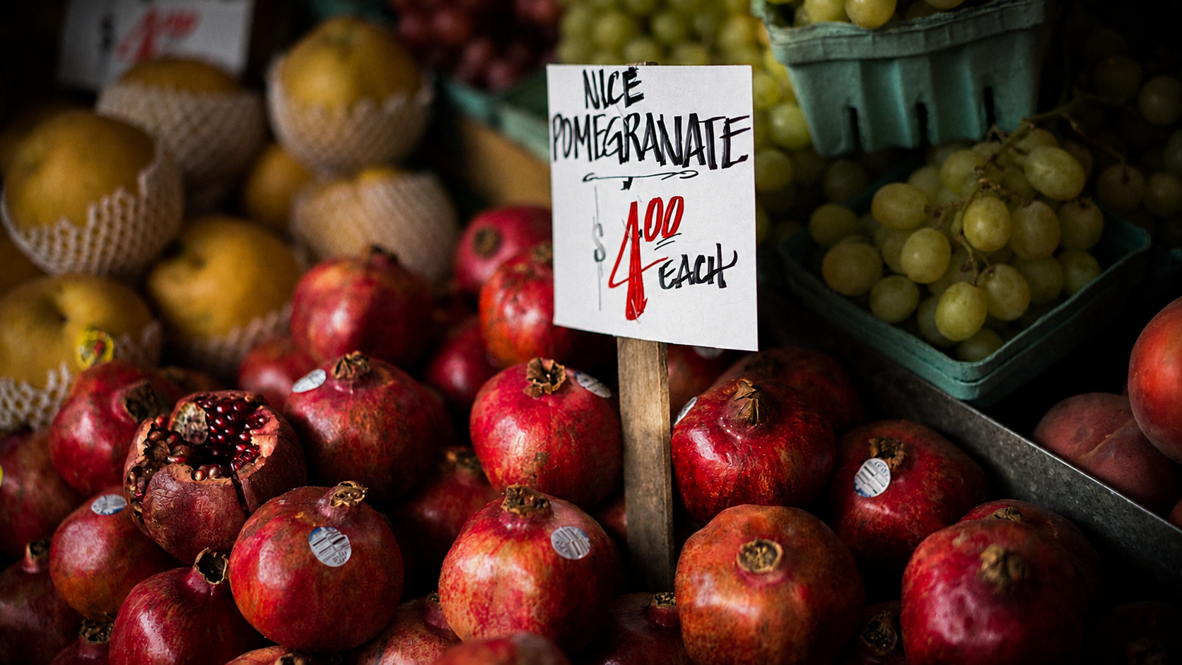 Fresh pomegranates displayed at a market with a sign indicating they are 'Nice Pomegranate' sold for $4 each, and green grapes in a green container on the side.