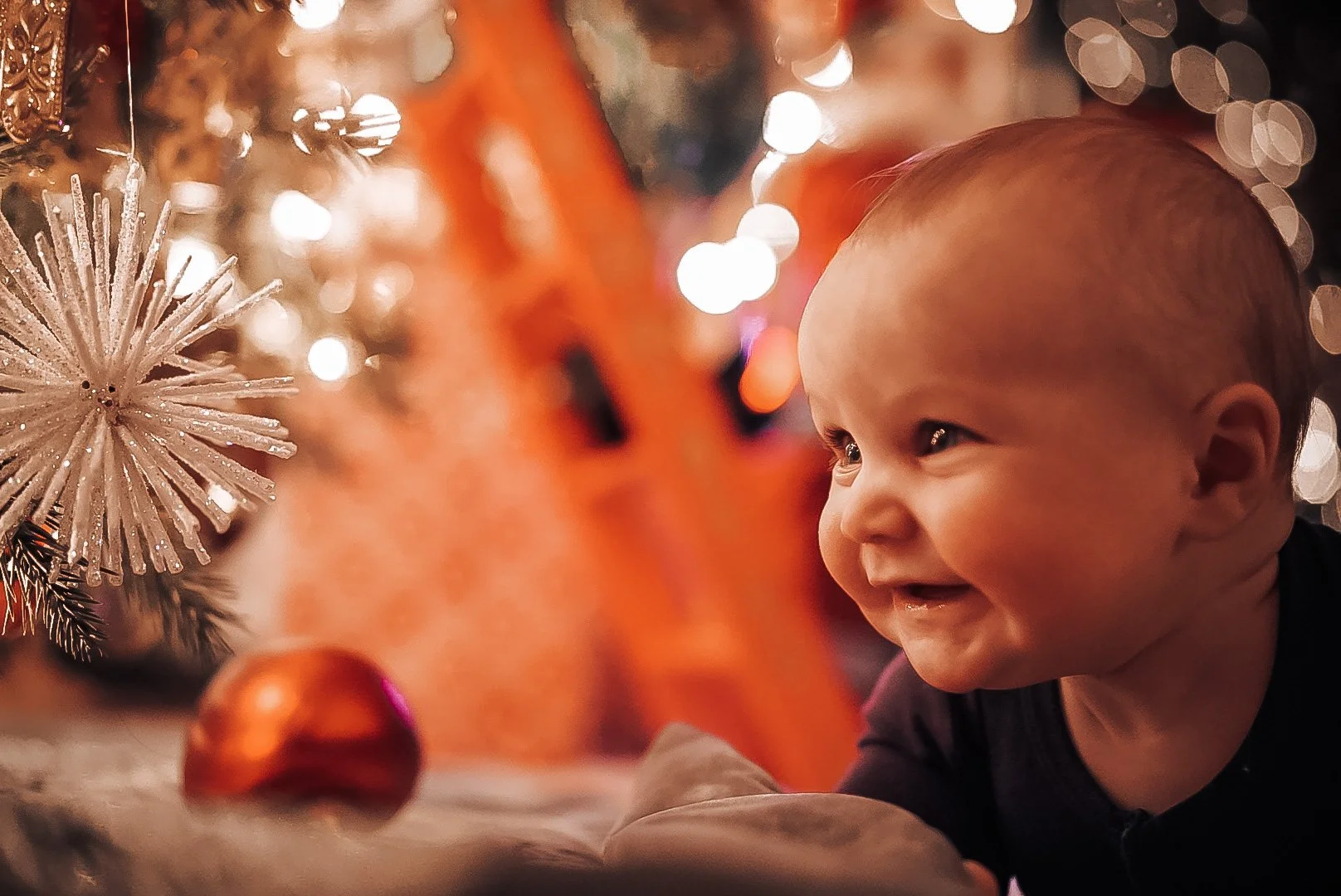 A baby with a big smile lying on the floor near a decorated Christmas tree with ornaments and bright lights.