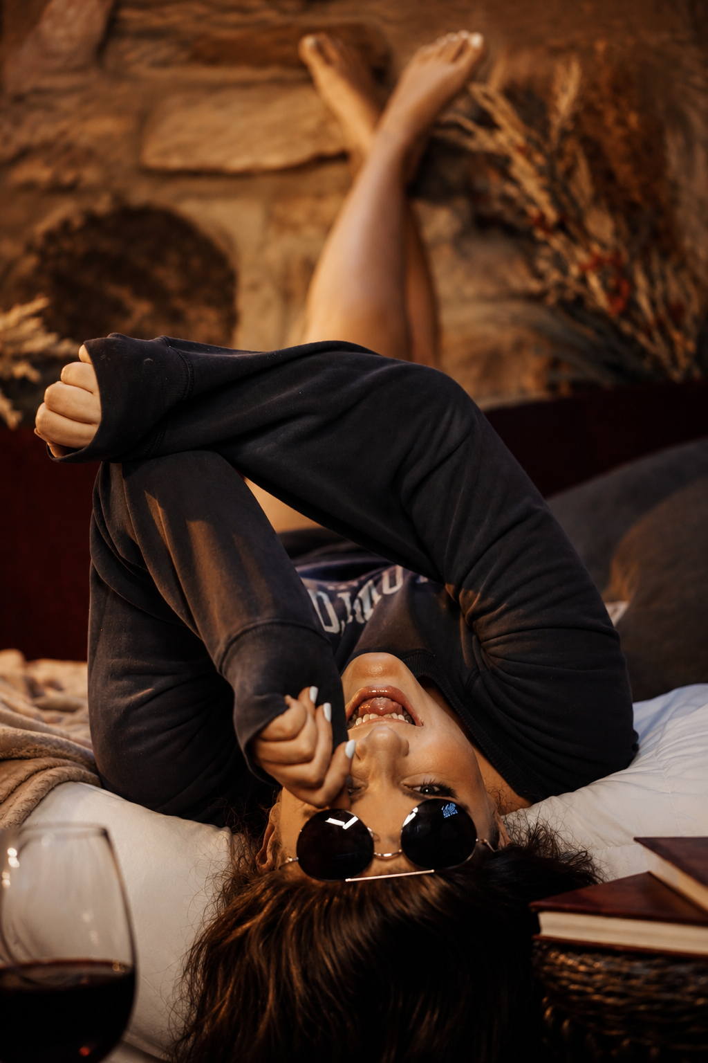 A woman lying on her back on a bed, wearing black sunglasses and a black hoodie, smiling and holding her hand near her face. Her head is propped up on a white pillow, with a glass of red wine and some books on a side table nearby. There is a large, t