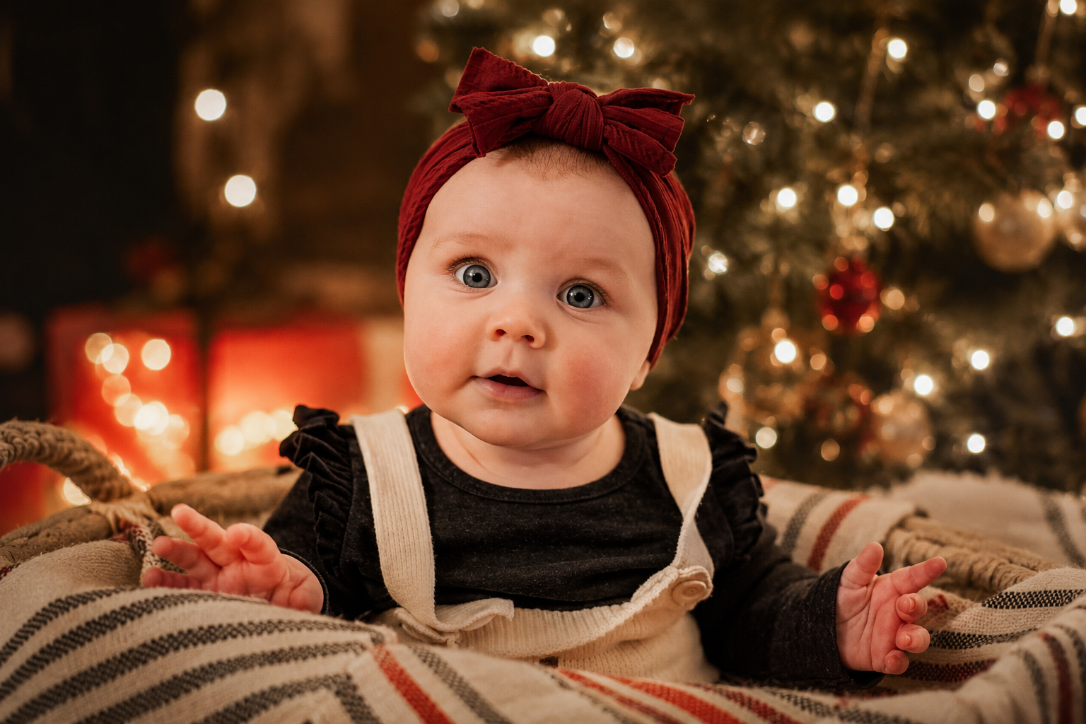 A baby with blue eyes wearing a red headband and dark shirt, sitting in front of a decorated Christmas tree with lights and ornaments.