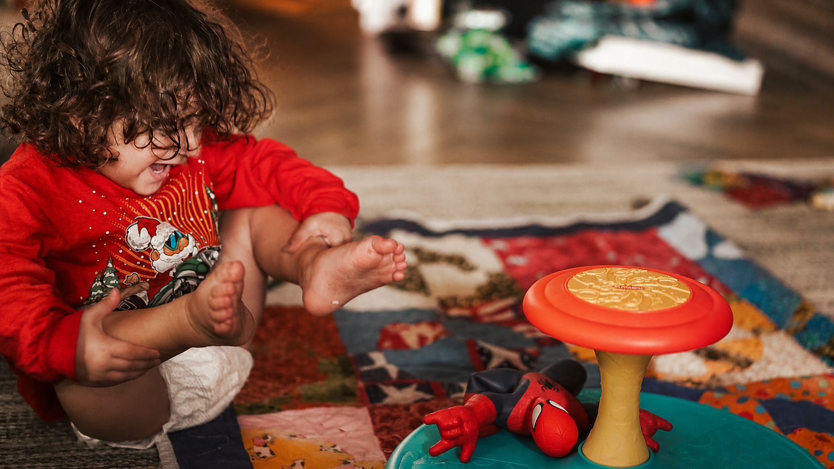 A young child with curly hair wearing a red Christmas-themed shirt playing on a colorful quilt with a Spider-Man toy and a spinning game.