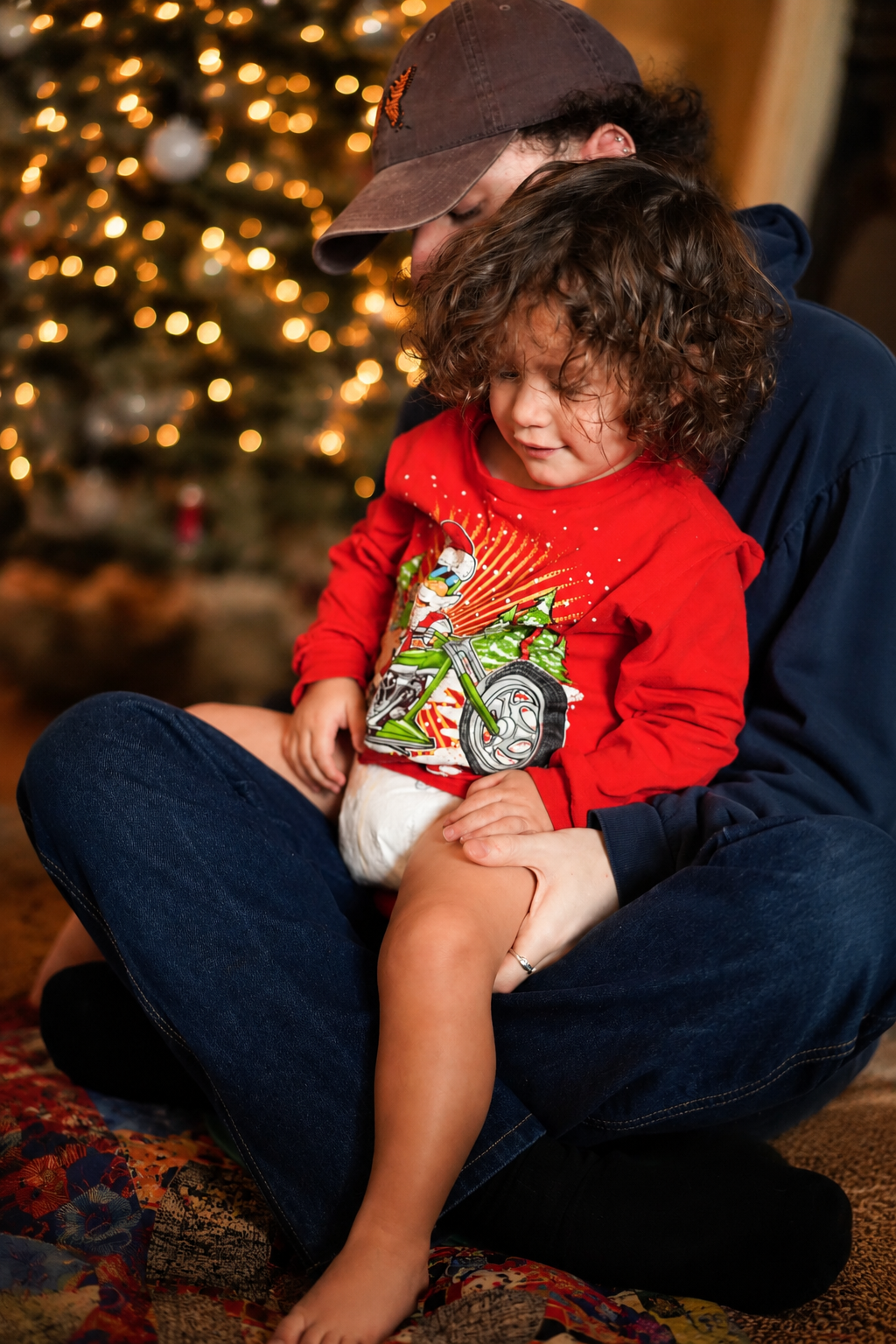 A man and a young child sitting on the floor in front of a decorated Christmas tree with blurred lights in the background. The adult is wearing a dark hoodie and a cap, while the child is wearing a red Christmas-themed shirt and sitting on the adult'
