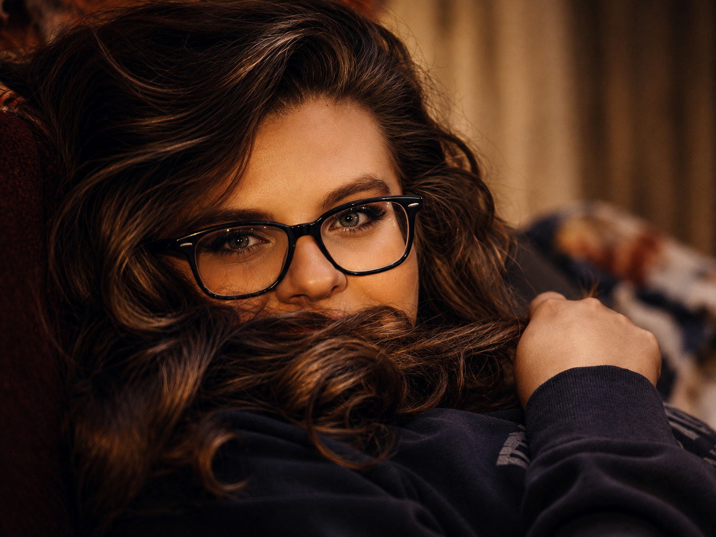 A woman with curly brown hair and glasses lying on a couch, looking at the camera.