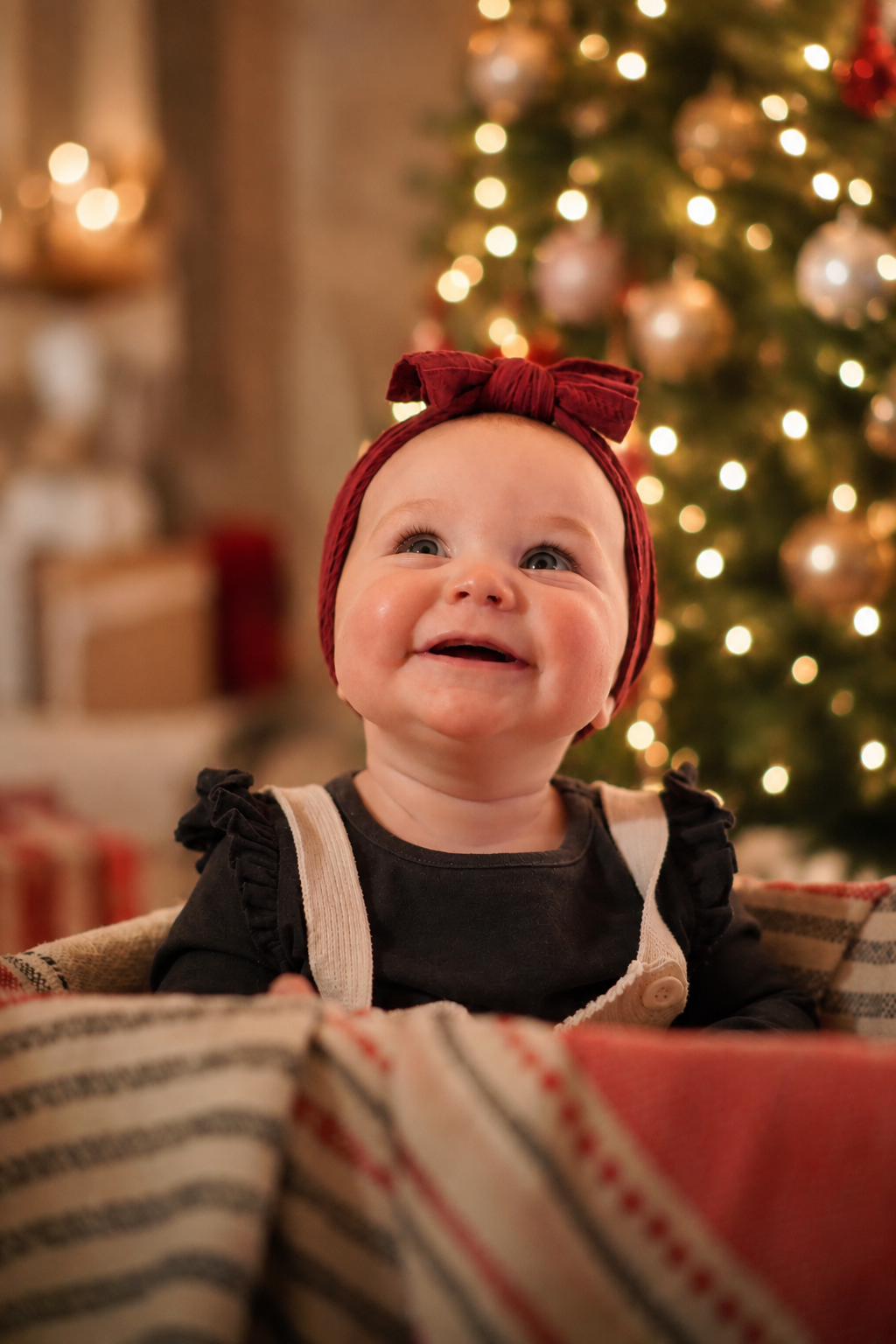 A smiling baby girl with blue eyes wearing a red headband with a bow, sitting in a festive environment with a decorated Christmas tree in the background.