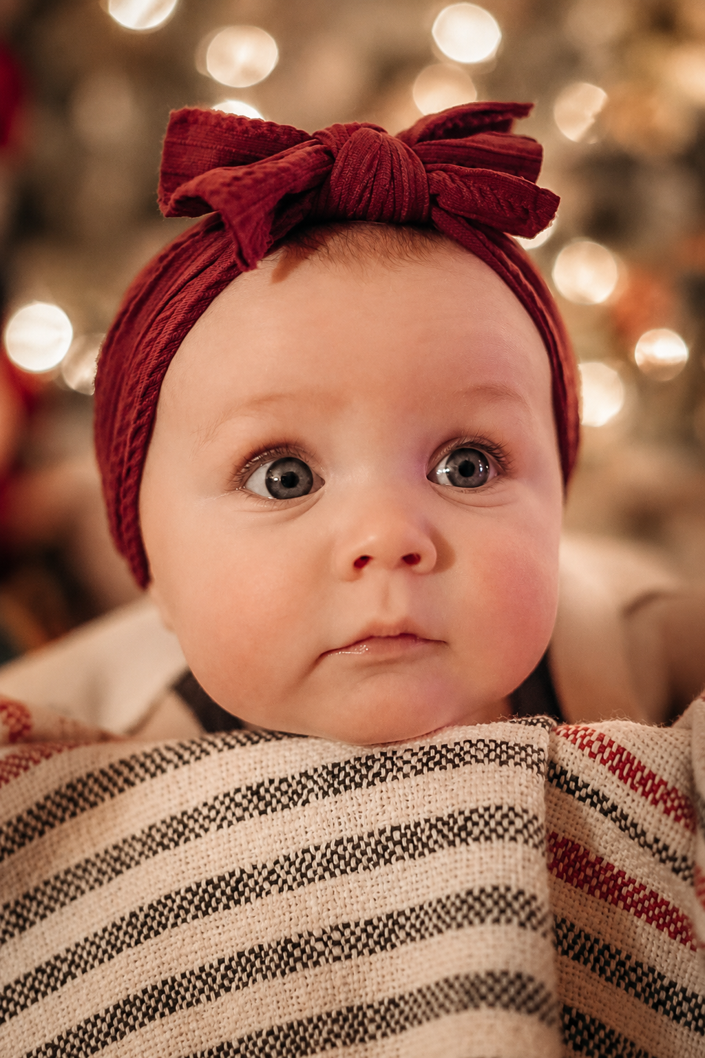 A close-up of a baby girl with big blue eyes, wearing a red headband with a bow and a black and white striped shirt, in front of a blurred background with Christmas lights.
