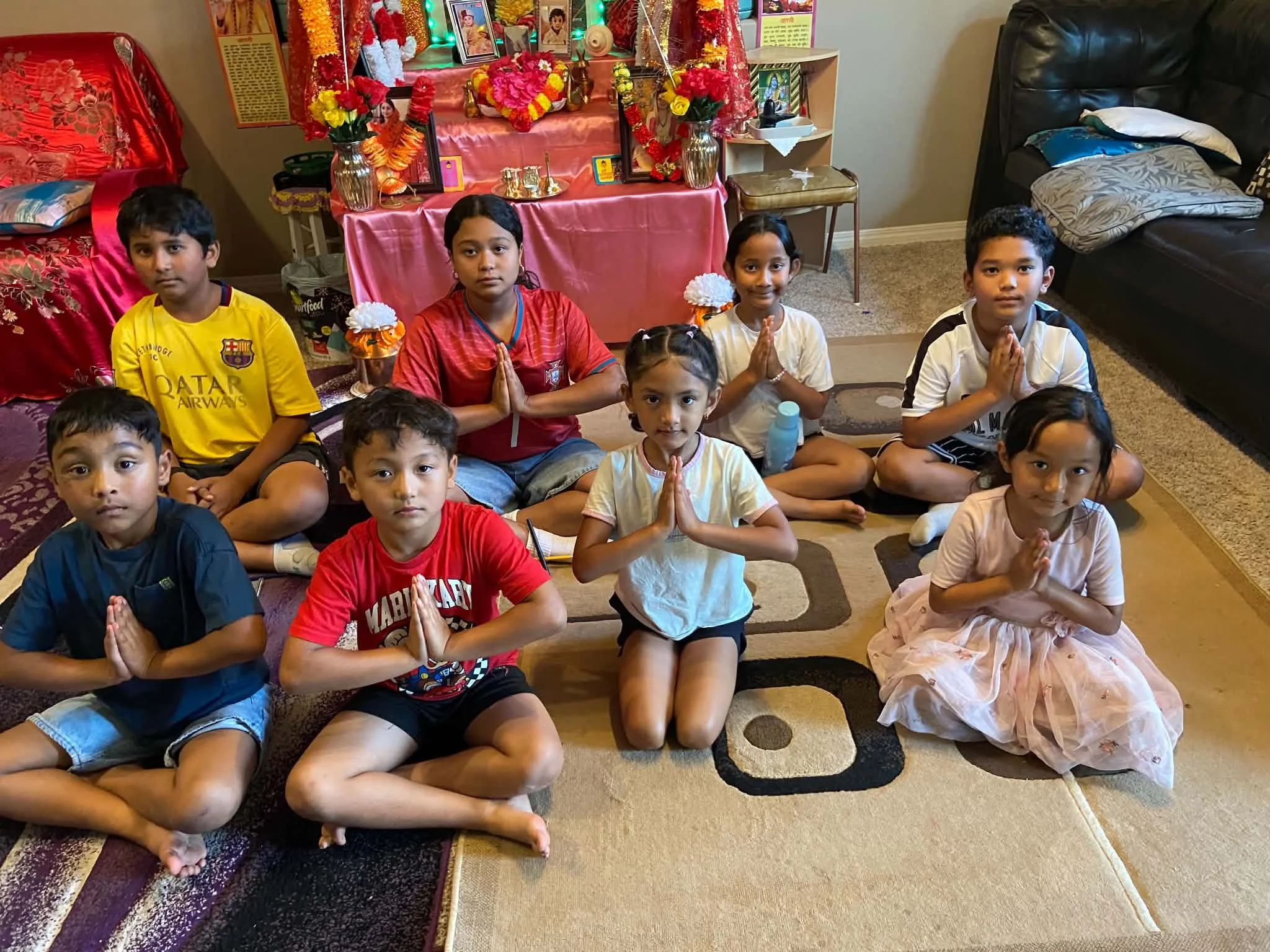 A group of young children sitting cross-legged on the floor, praying with their hands pressed together in front of a decorated altar. The altar is adorned with flowers, framed photographs, and religious items, indicating a Hindu religious or cultural ceremony.
