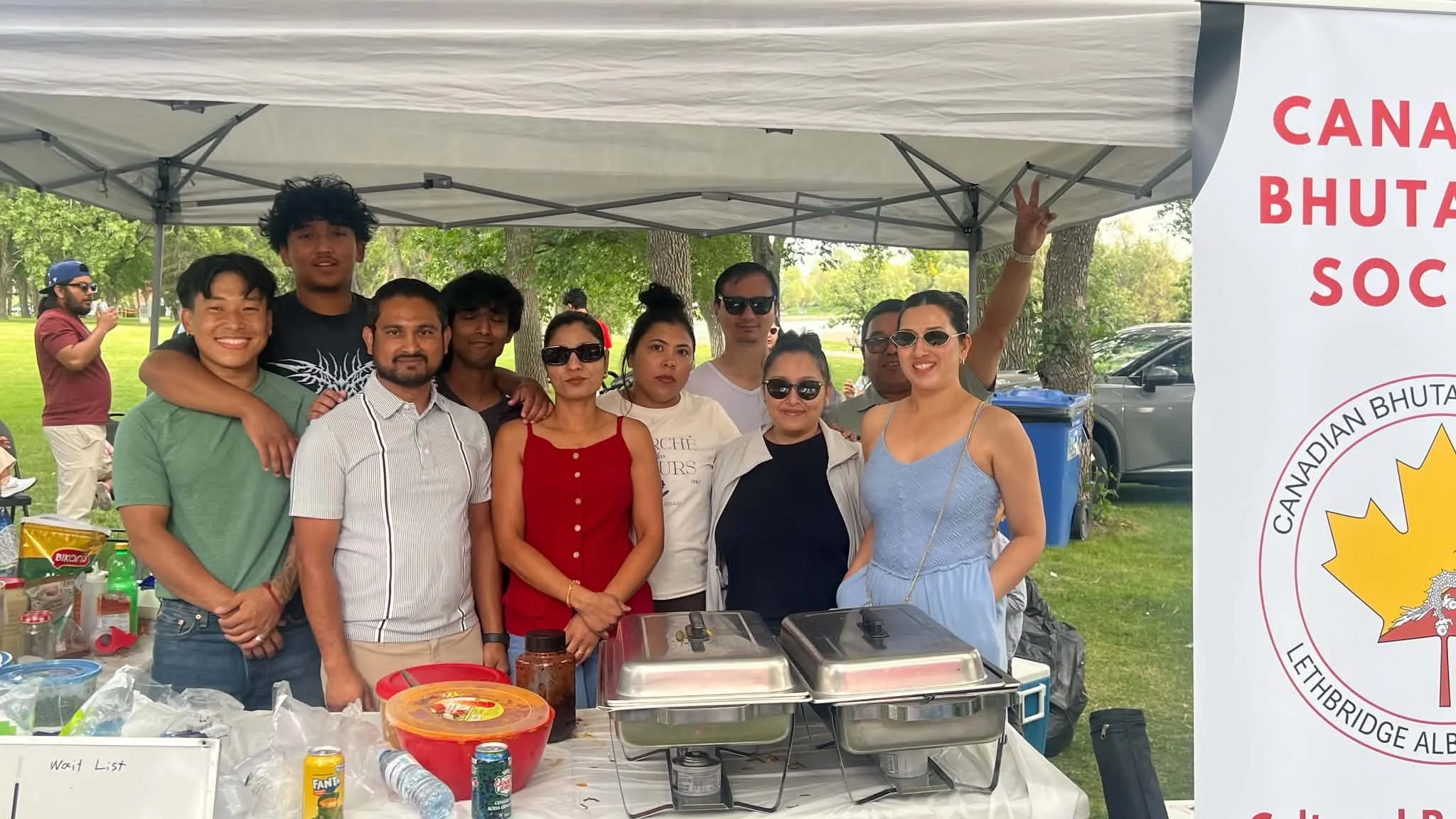 Group of people standing under a canopy tent outdoors, smiling, with a banner to the right that reads 'CANADIAN BHUTAN SOC' and part of a logo visible. There are trees in the background and a table with food and drinks in front of them.