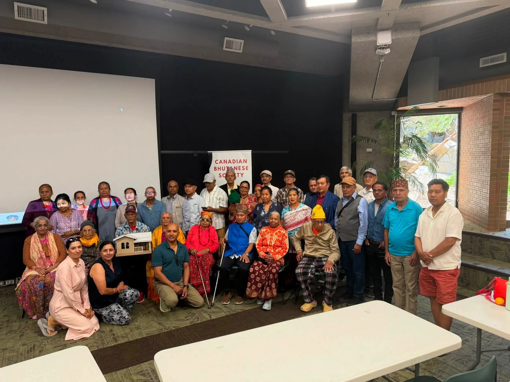 A large group of people gathered together in an indoor setting, likely for a community or cultural event. The group includes men and women of various ages, some dressed in traditional South Asian attire, while others are in casual clothing. There is a sign that reads "Canadian Bhutanese Society" in the background. The room has high ceilings, large windows with greenery visible outside, and some tables in the foreground.