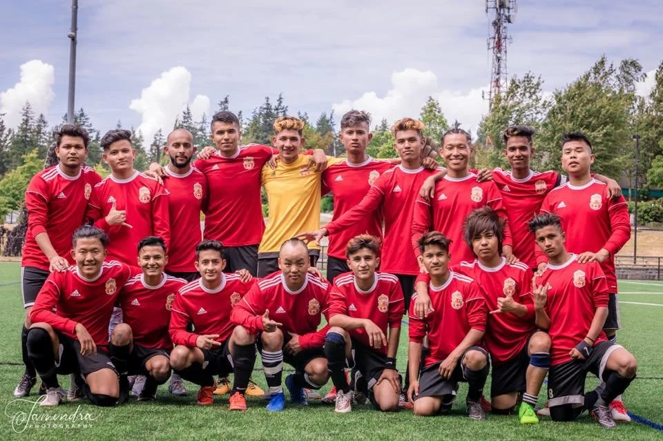 Group of young male soccer players posing on a field, wearing red jerseys with a crest, with one in a yellow goalkeeper jersey, outdoor setting with trees and cloudy sky.