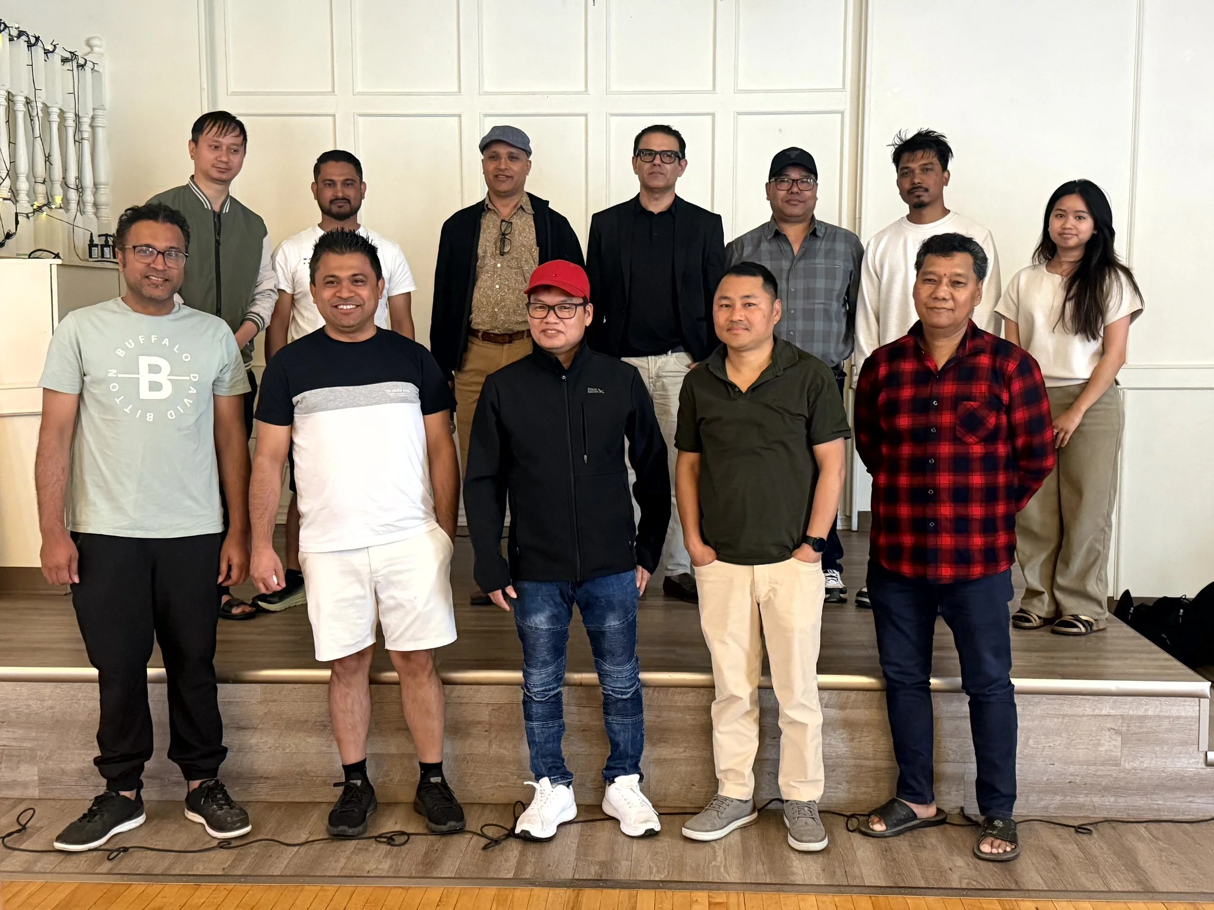 Group of fifteen people standing indoors for a photo, some smiling and some not, with a white paneled wall in the background.