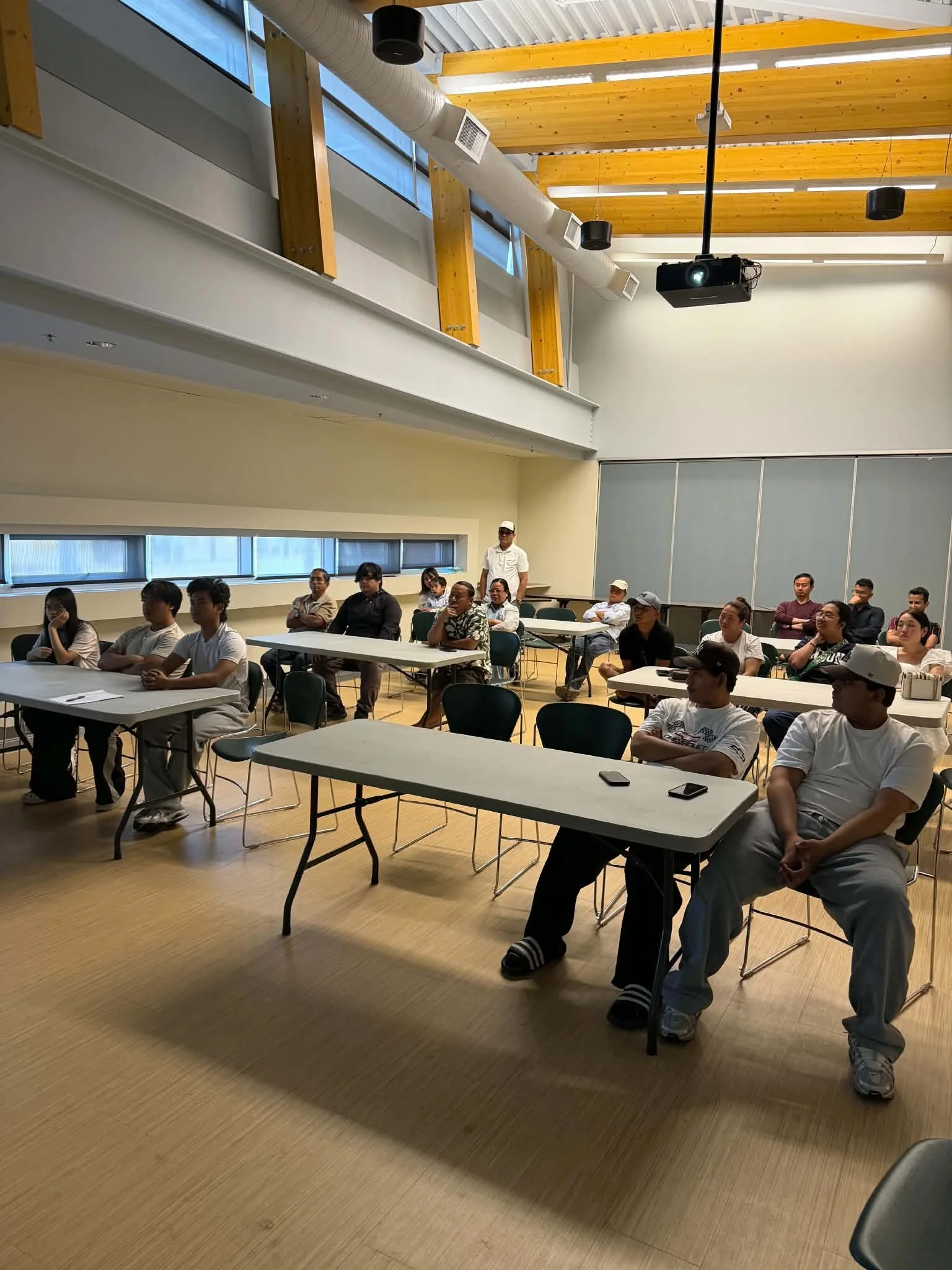 People attending a presentation or lecture in a classroom or conference room with large windows and wooden ceiling beams.